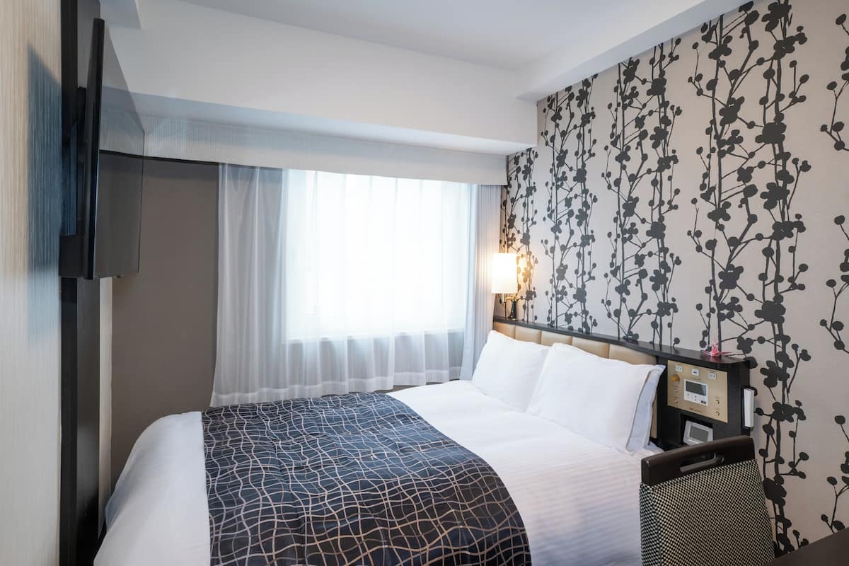 A hotel room with a bed, a TV, a bedside table with a lamp, and a wall with a floral pattern.
