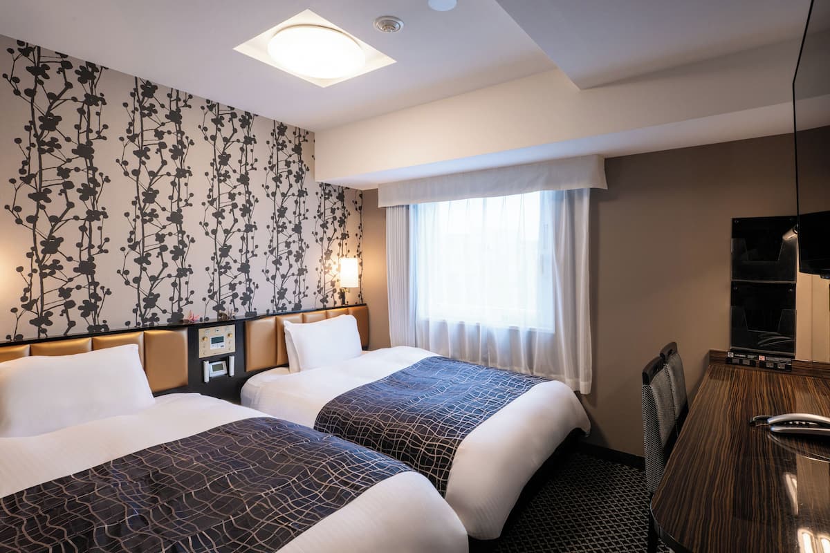 A hotel room with two beds, a desk, a TV, and a window with curtains.