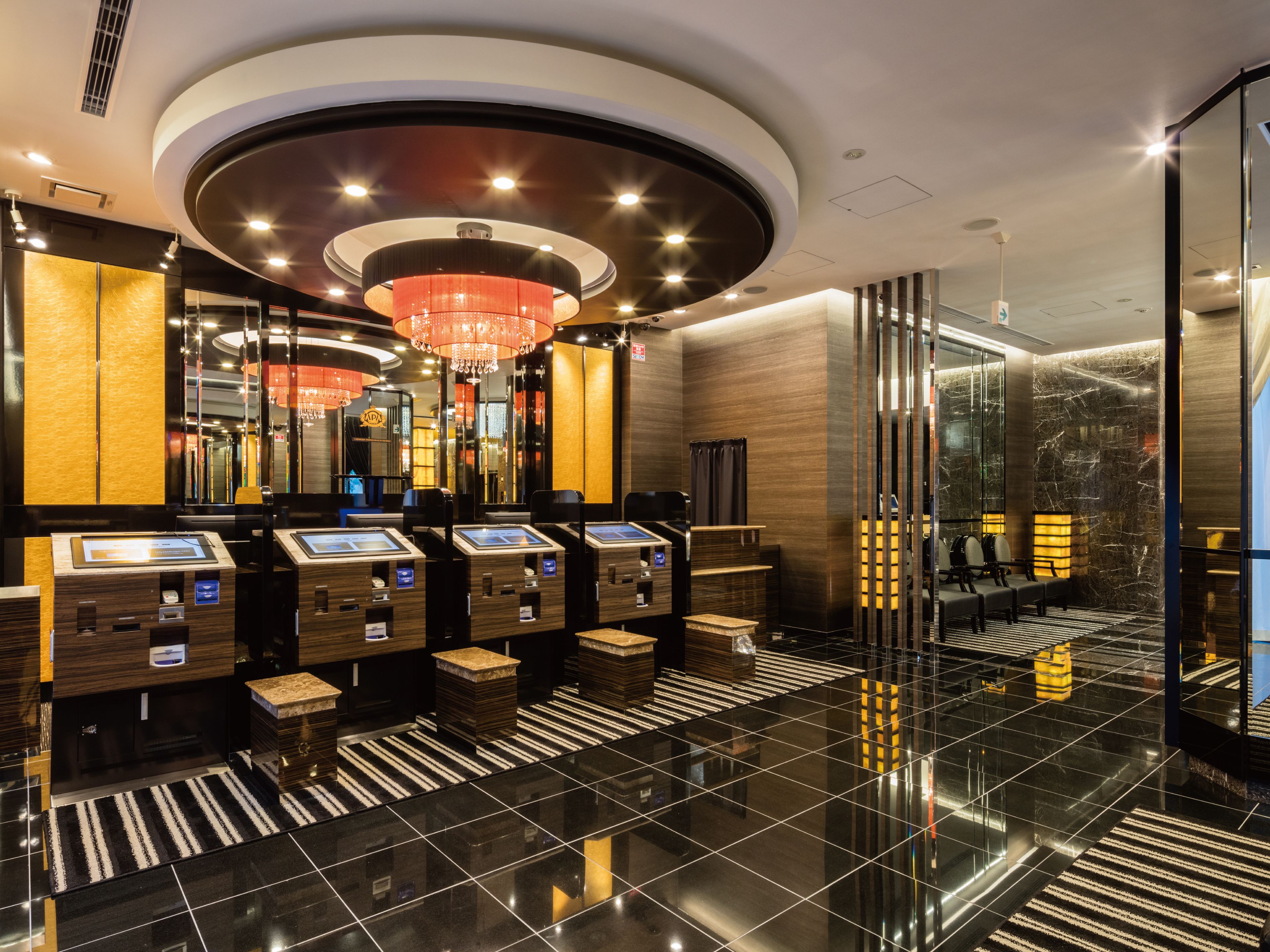 A modern hotel lobby with a central chandelier, check-in kiosks, and a reflective floor.