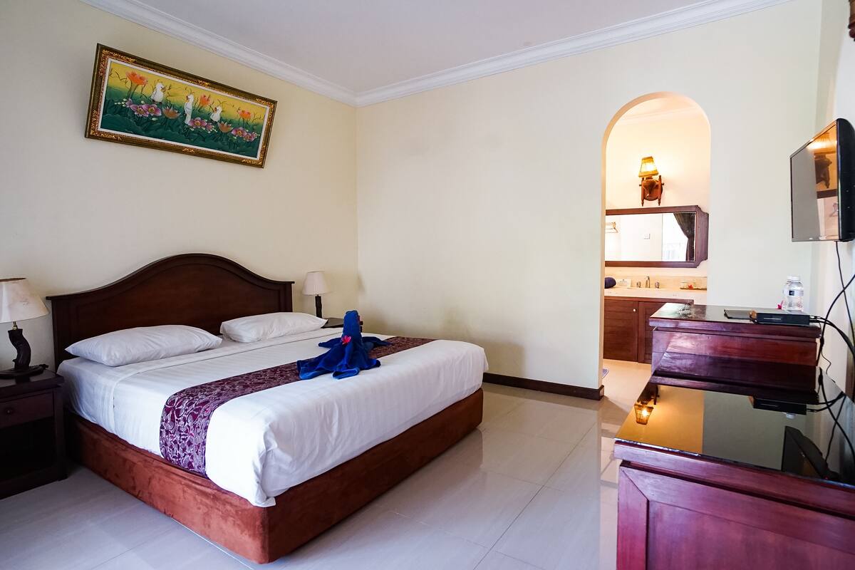 Hotelkamer met een groot bed, een houten hoofdeinde, een schilderij aan de muur, een bureau met een lamp en uitzicht op de keuken.