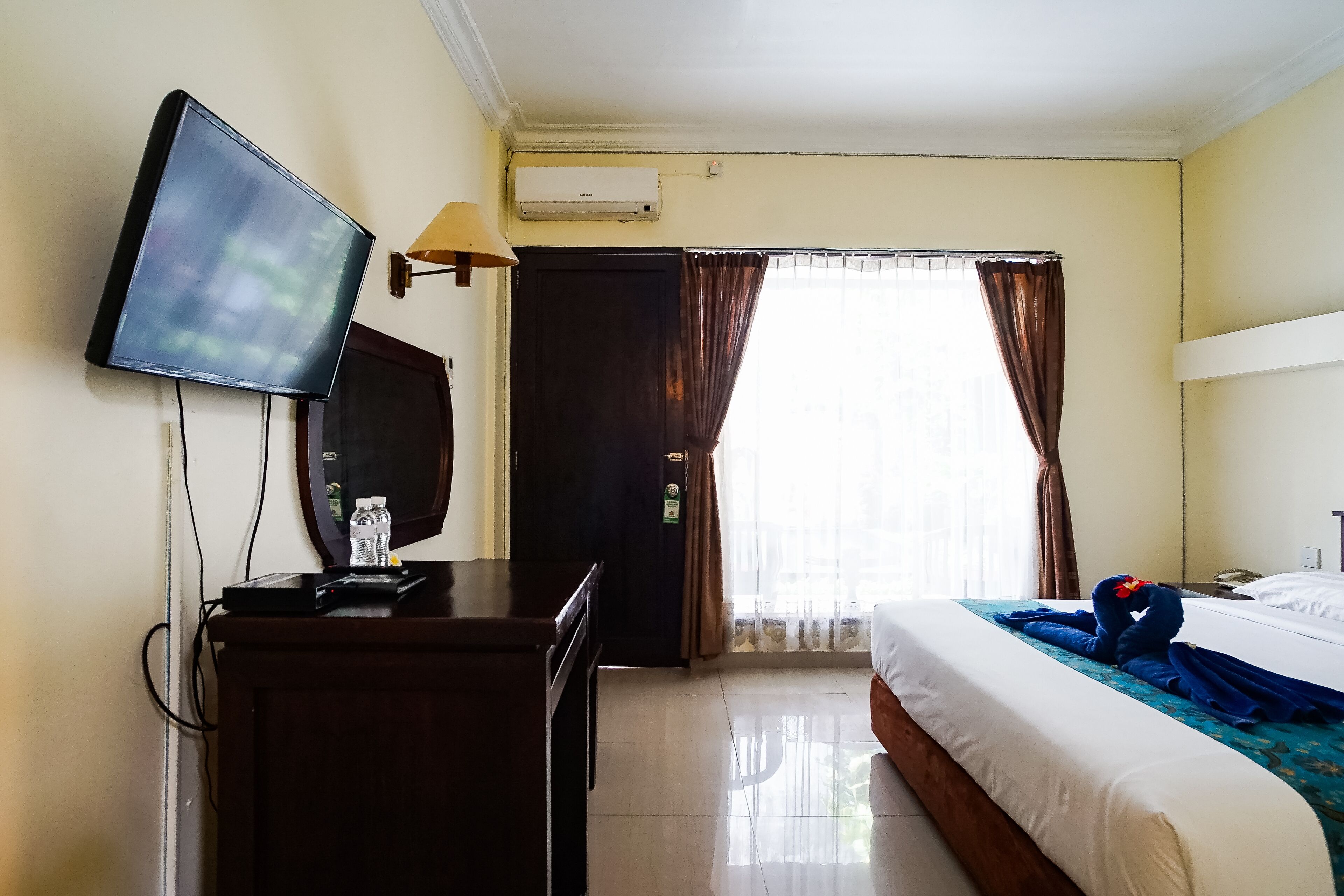 A hotel room with a bed, a desk with a flat-screen TV, a lamp, and a window with curtains.
