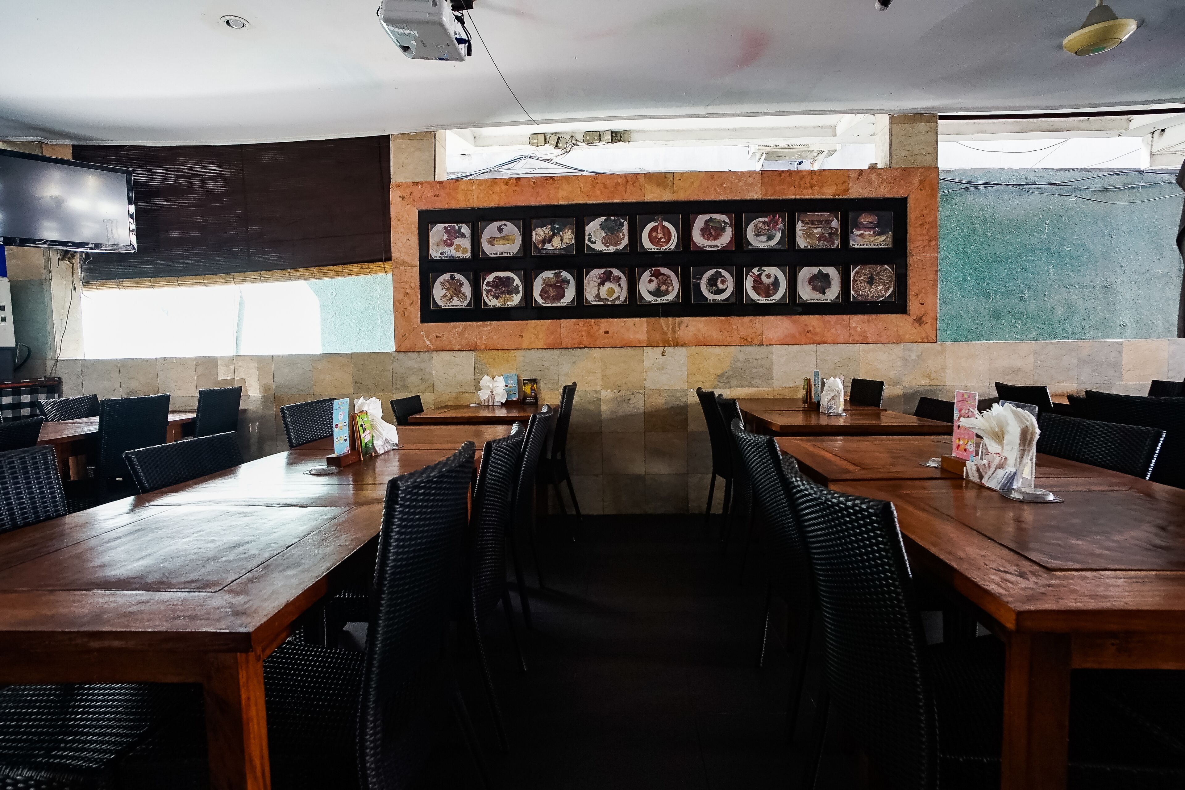 A restaurant with multiple wooden tables and black chairs, a wall-mounted menu board, and a large window.
