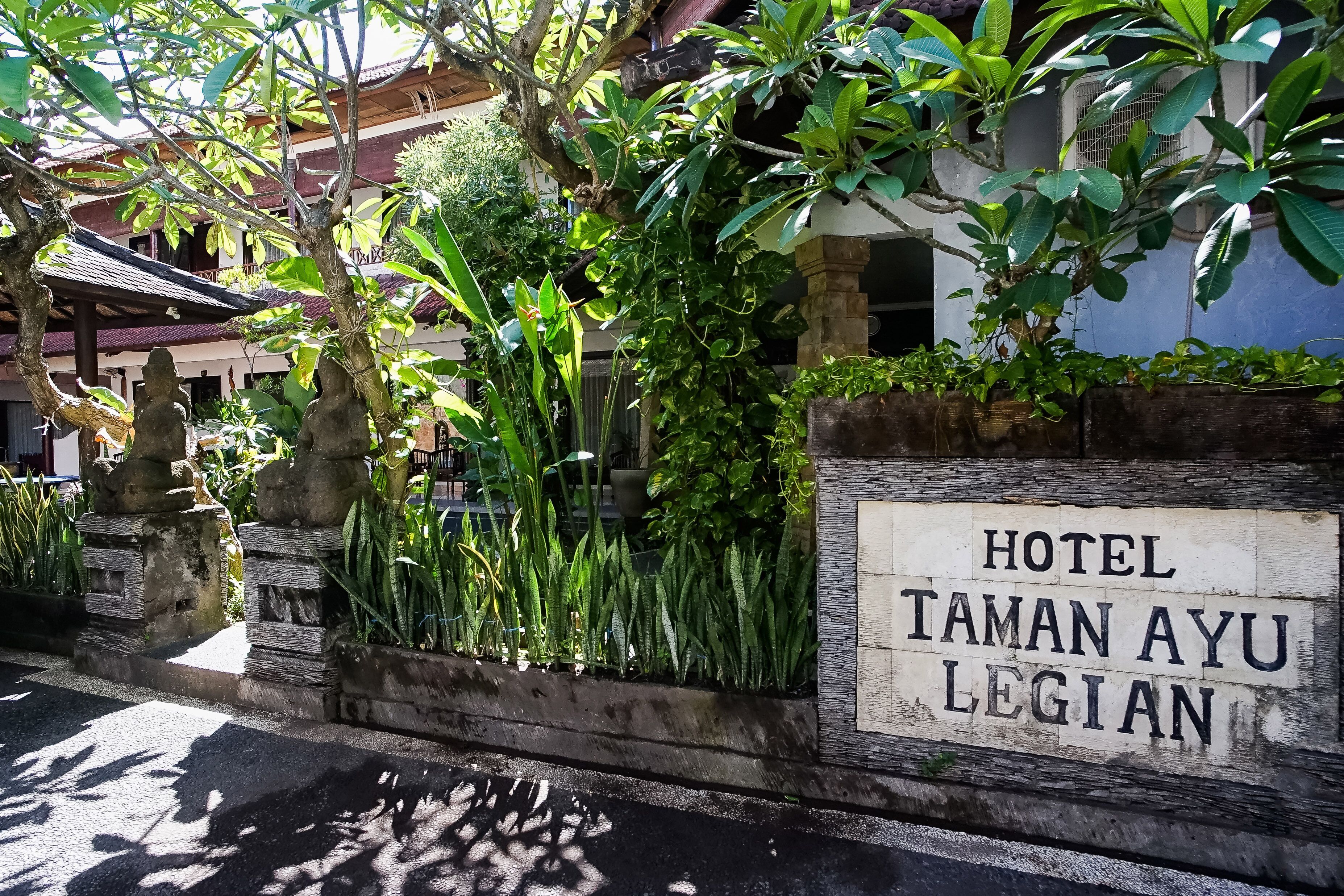 A sign for Hotel Taman Ayu Regency with a stone wall and lush greenery.