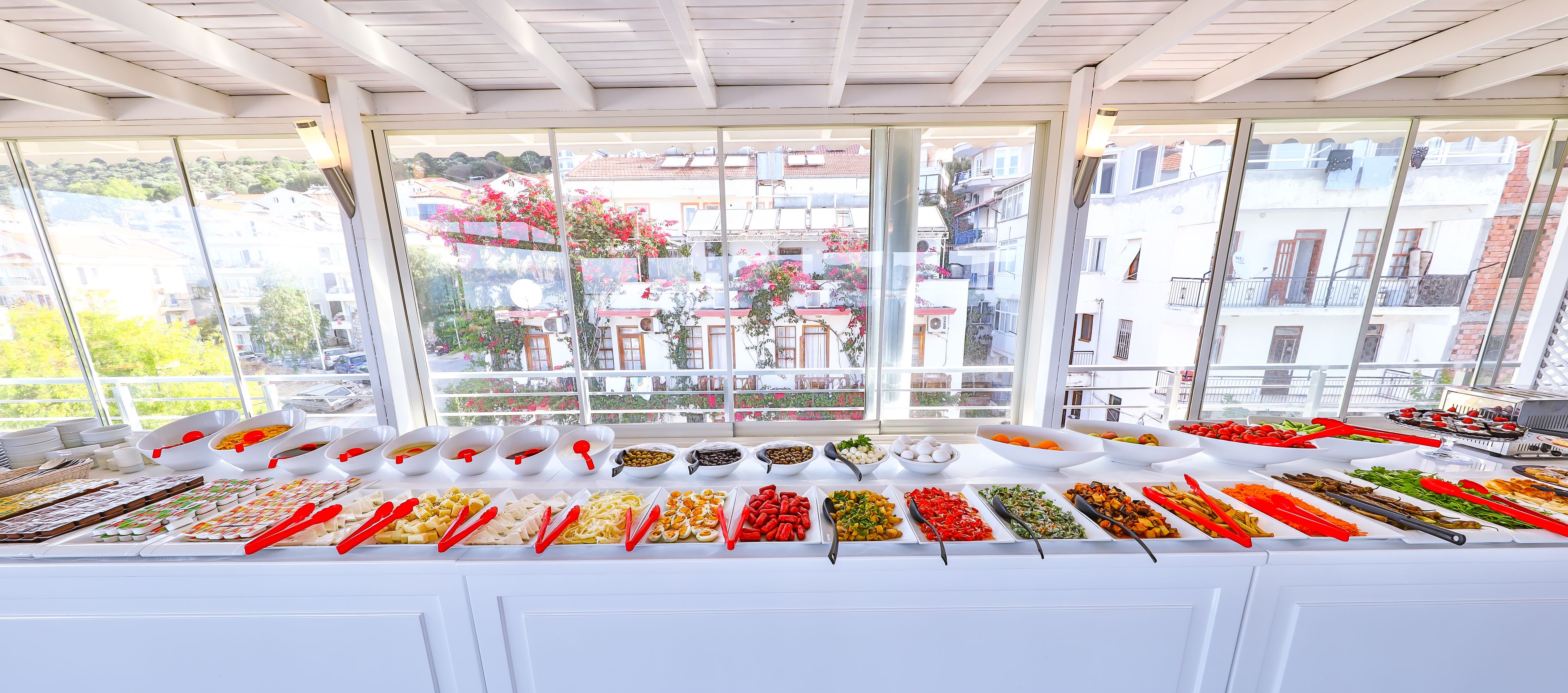 A buffet with various food items and utensils, set against a backdrop of a balcony with a view of buildings and greenery.