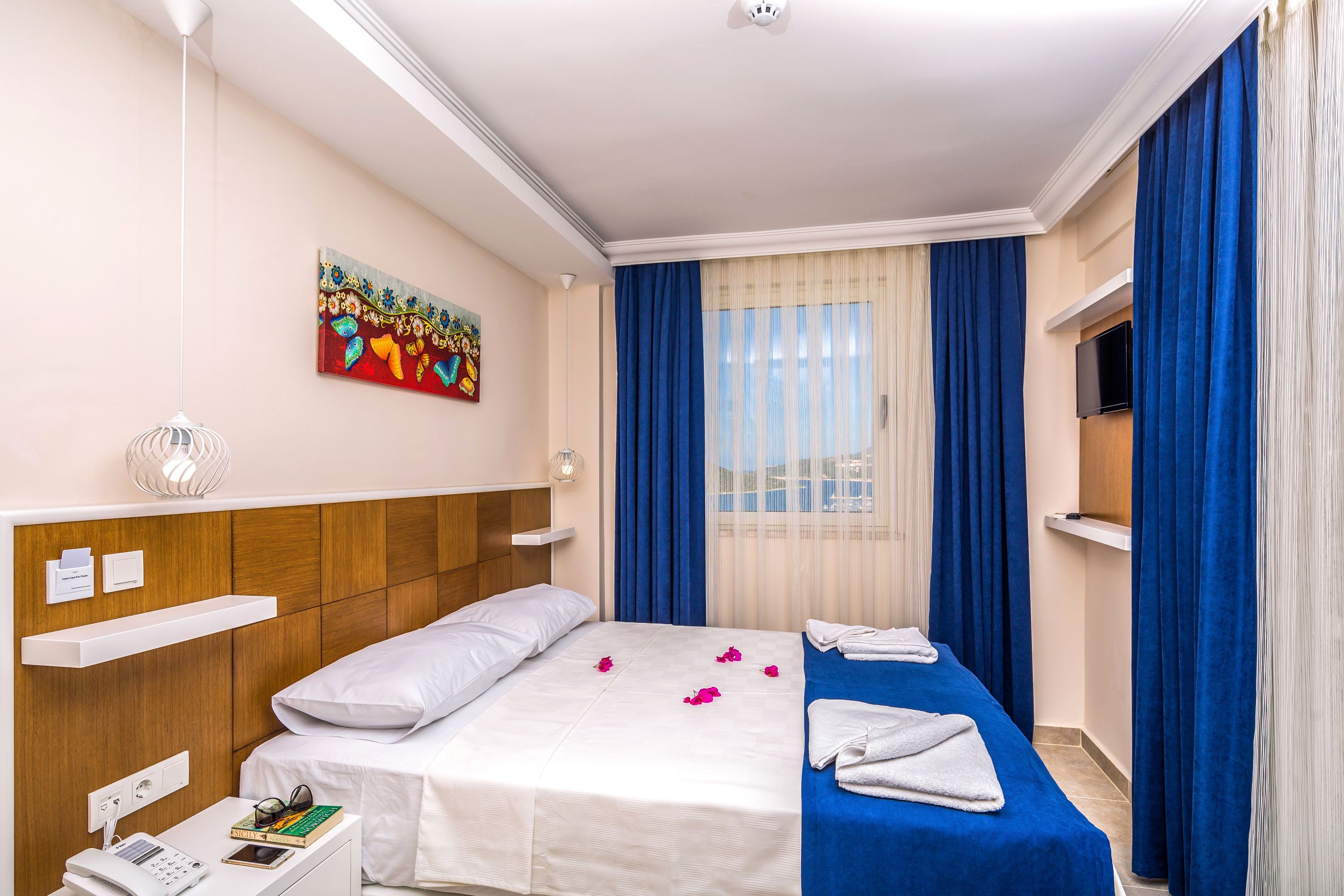 A hotel room with a bed, a bedside table with a phone and books, a painting on the wall, and a TV on a stand.