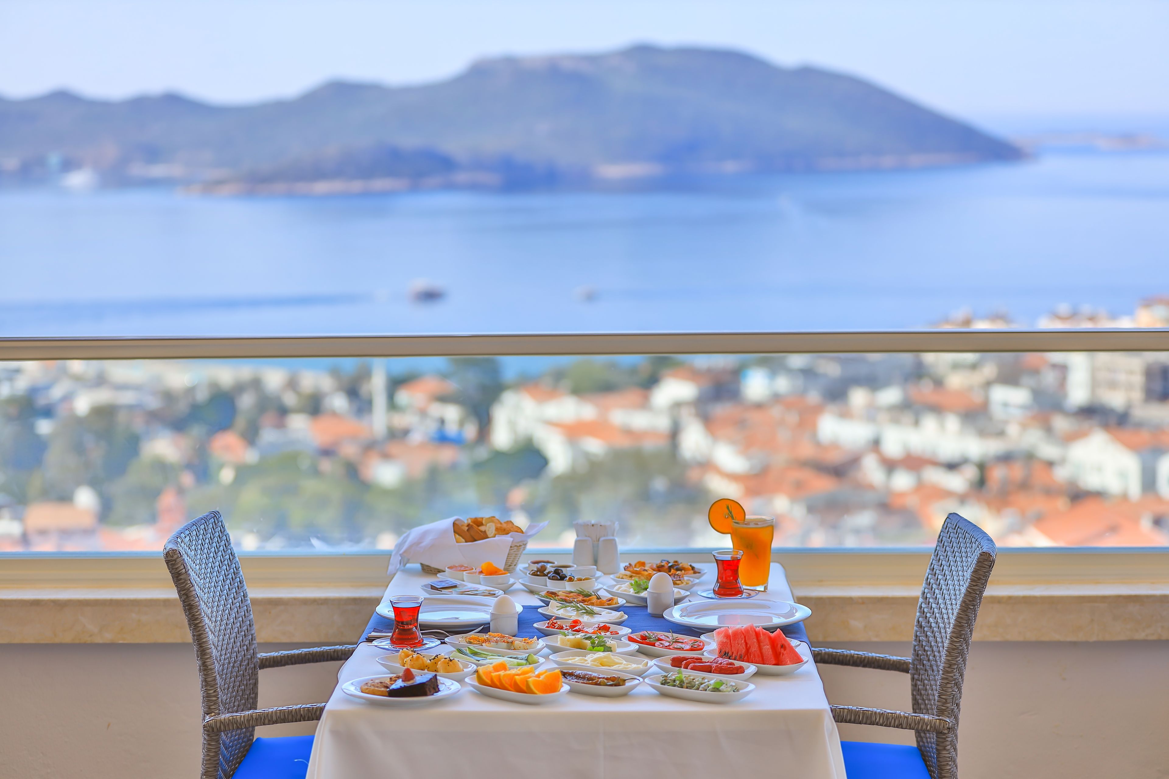 Una mesa dispuesta con varios platos y un vaso de jugo de naranja, con vistas a una ciudad costera con edificios y una montaña al fondo.
