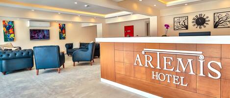A hotel reception area with a wooden reception desk, blue seating, and artwork on the walls.