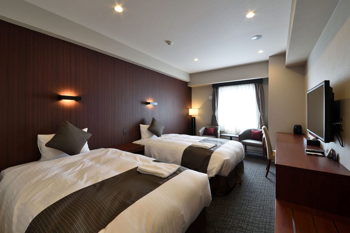 A hotel room with two beds, a desk, a chair, a TV, and a window with curtains.