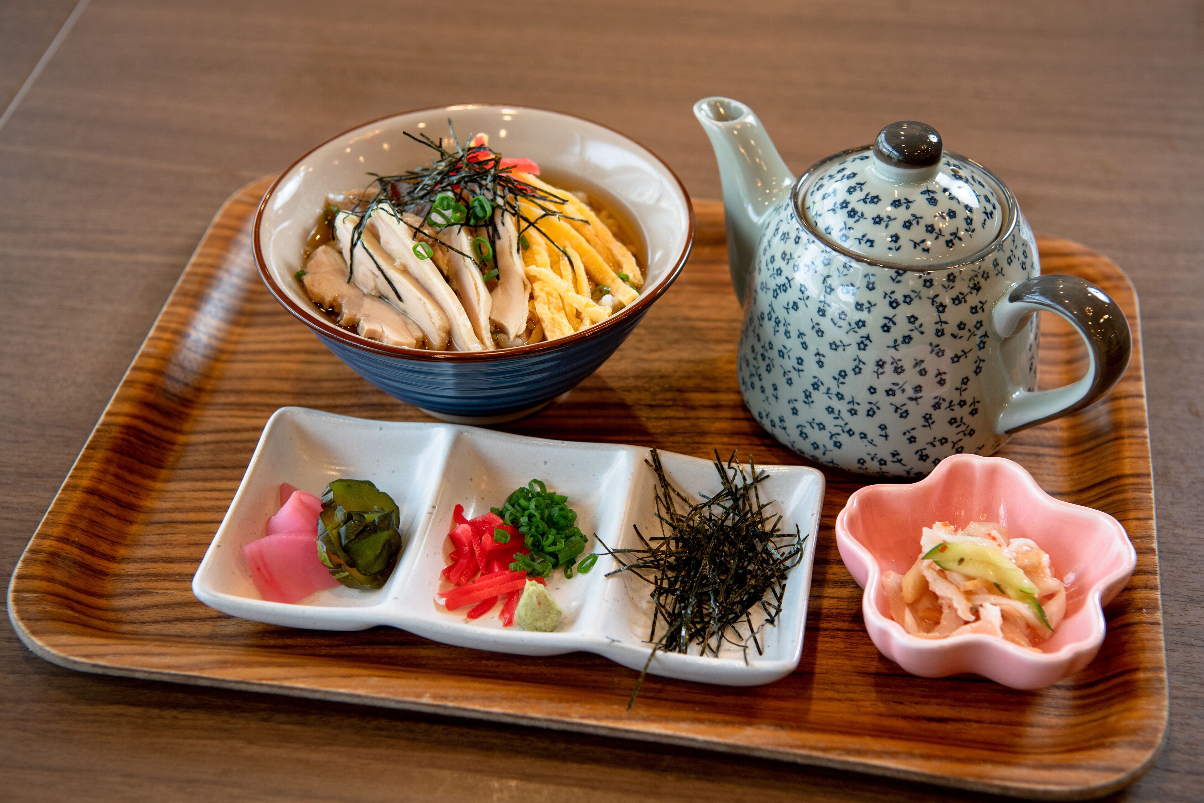 A wooden tray with a bowl of food, a teapot, and small dishes of condiments.