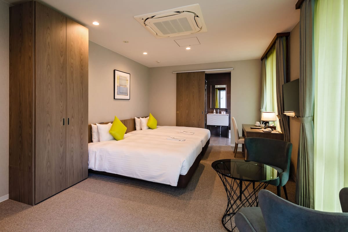 A hotel room with a large bed, a desk with a chair, a small table, and a wardrobe.