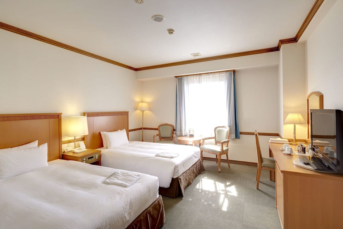 A hotel room with two beds, a desk, a chair, a television, and a window with curtains.