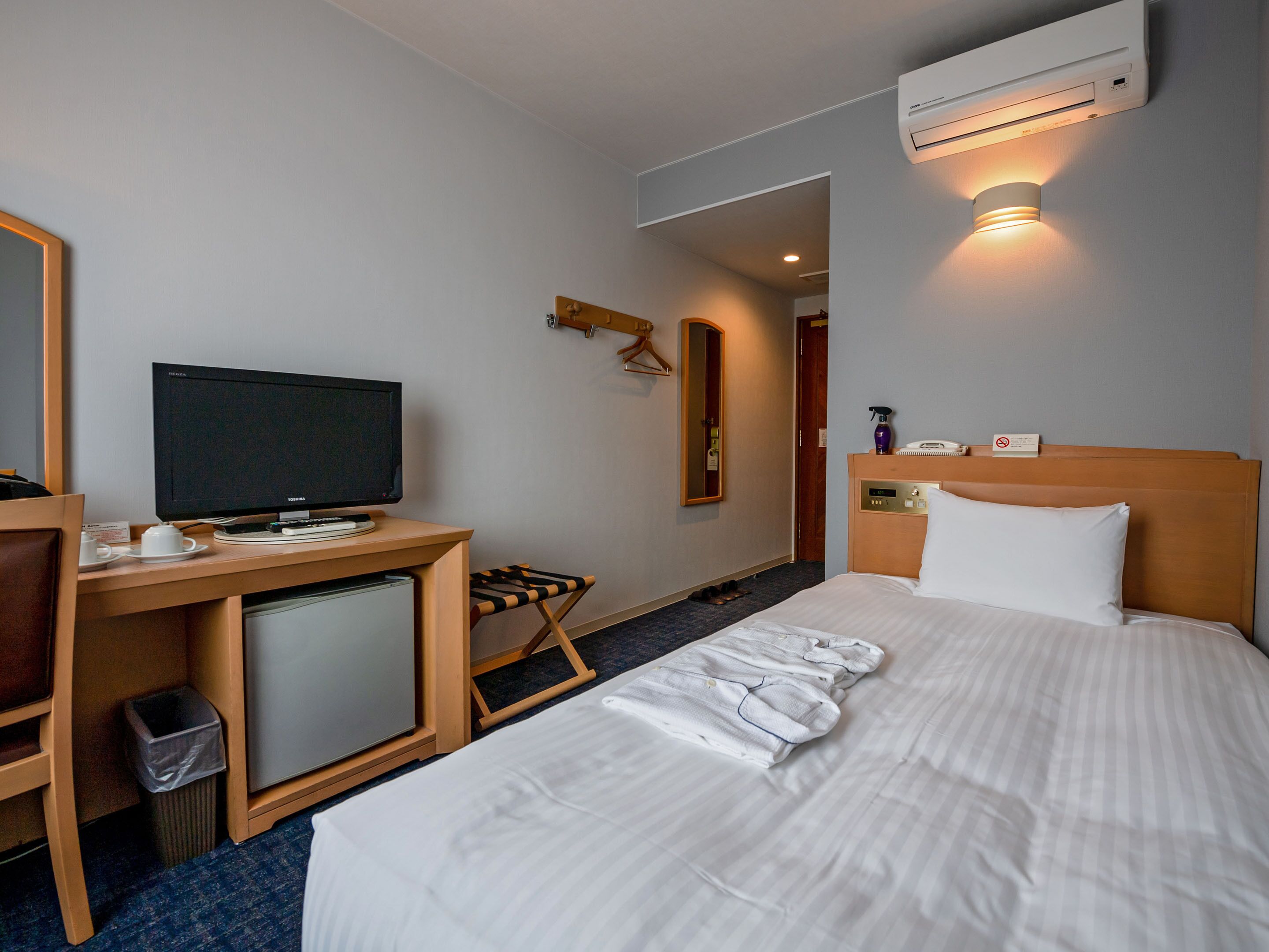 A hotel room with a bed, a desk with a TV, a mirror, and an air conditioning unit.