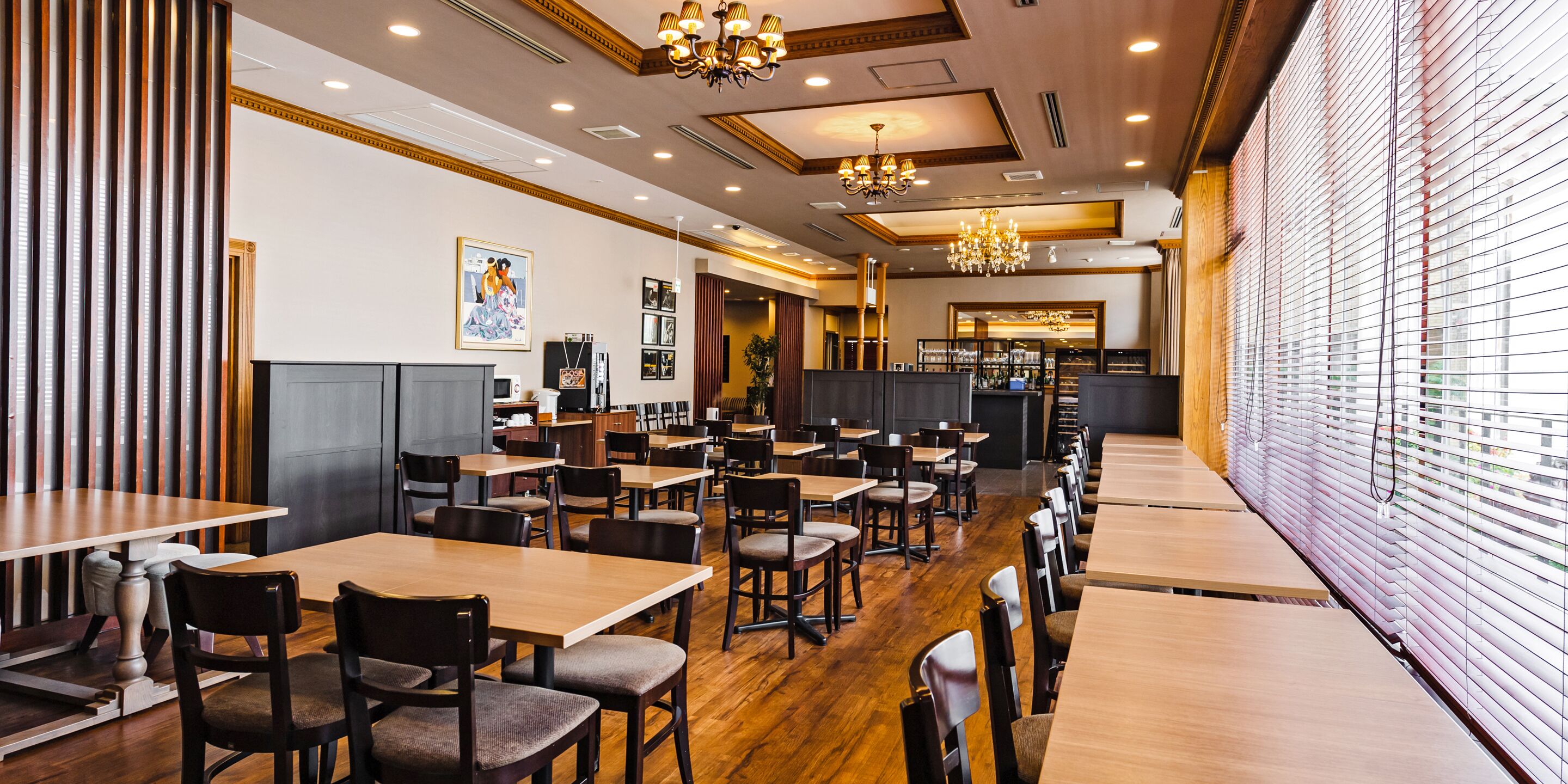 A restaurant with wooden tables and chairs, a chandelier, and a large window with blinds.