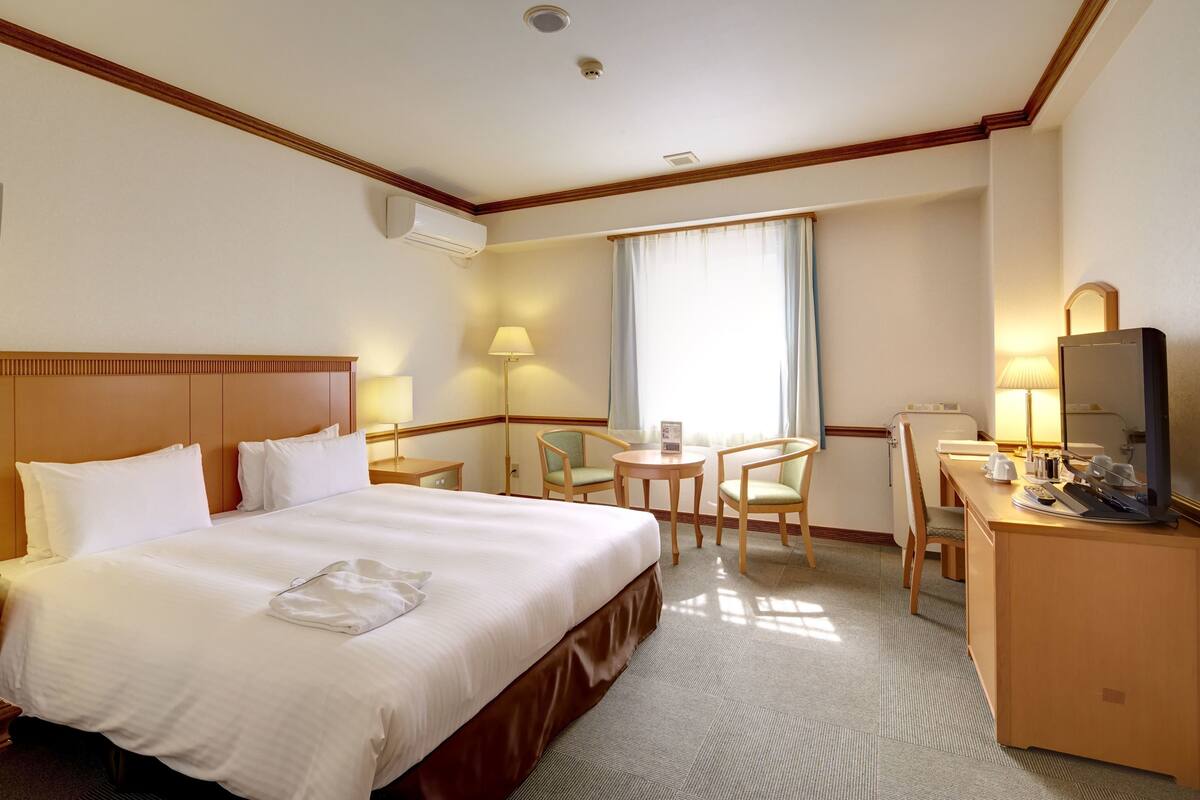 A hotel room with a large bed, a desk, two chairs, a television, and a window with curtains.