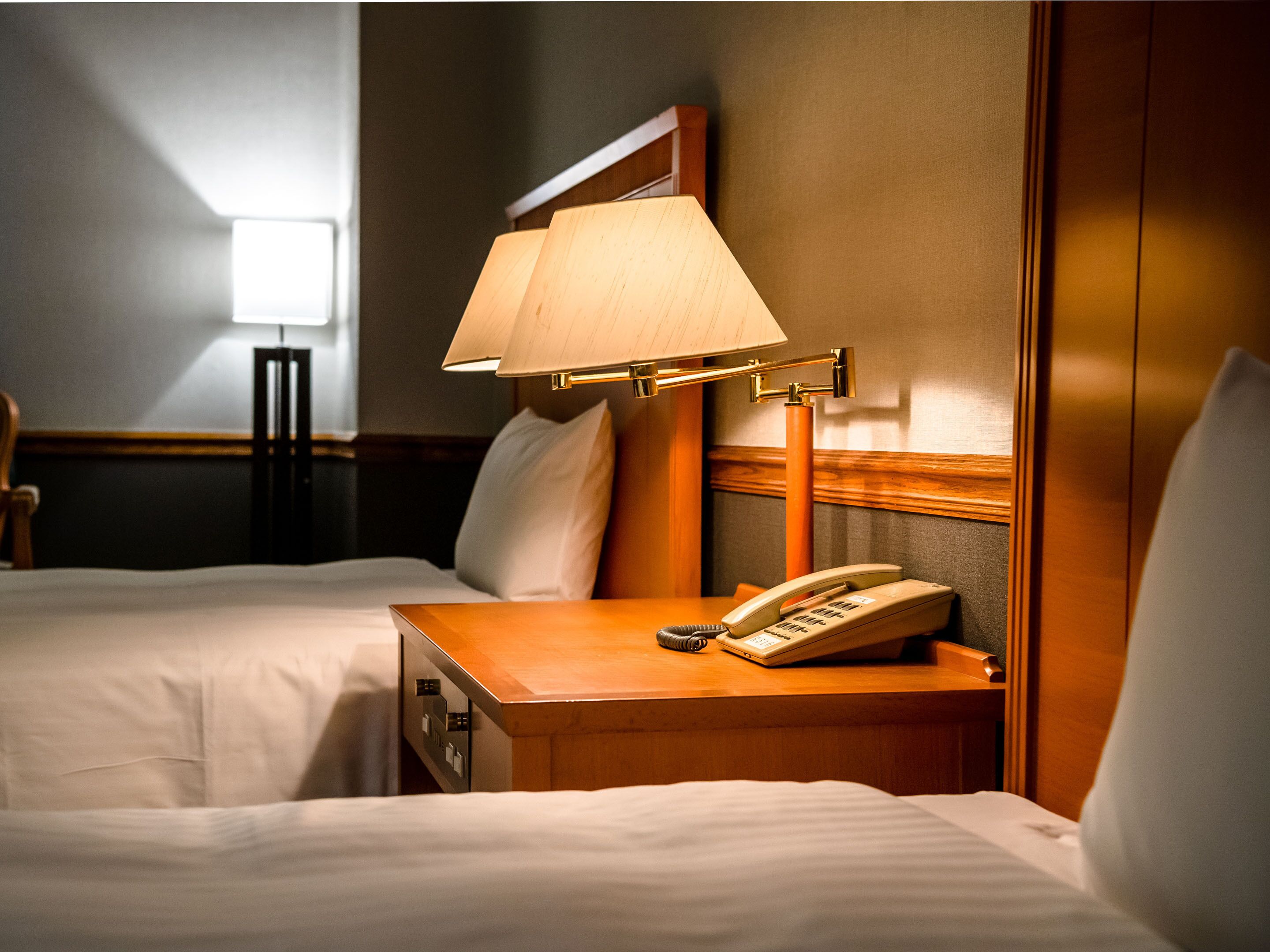 Hotellihuone, jossa on kaksi sänkyä, yöpöytä lampun kanssa, puhelin ja toinen pienempi pöytä.