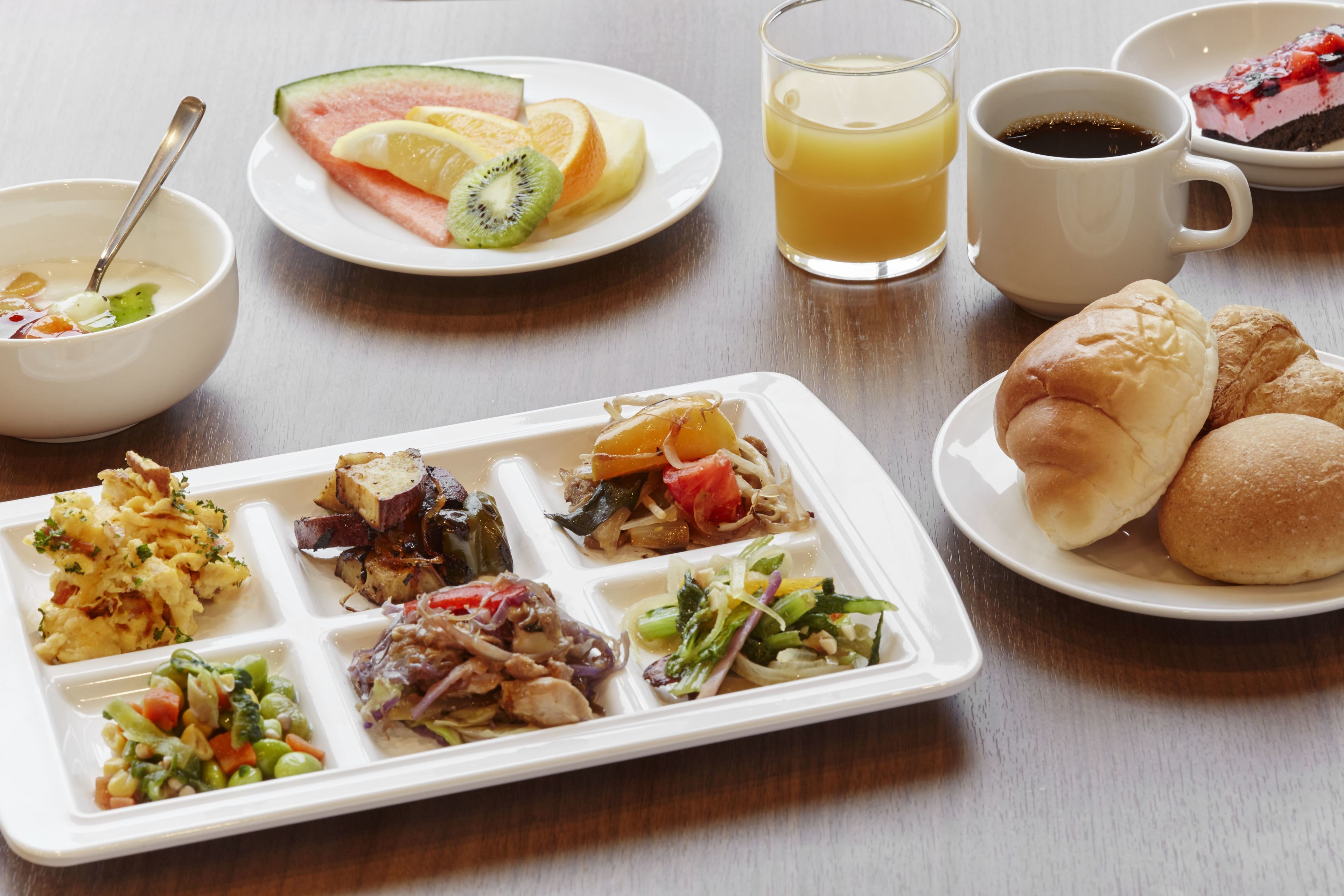 A meal with various dishes including a fruit plate, a glass of orange juice, a cup of coffee, and a plate with rolls and vegetables.
