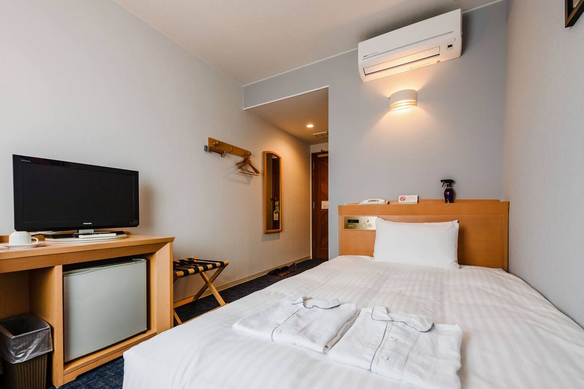 A hotel room with a bed, a desk with a television, a small refrigerator, a trash bin, and an air conditioning unit.