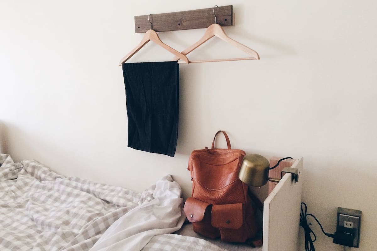 Un dormitorio con una cama, un perchero, un bolso de cuero marrón y una mesita de noche con una lámpara e interruptos.