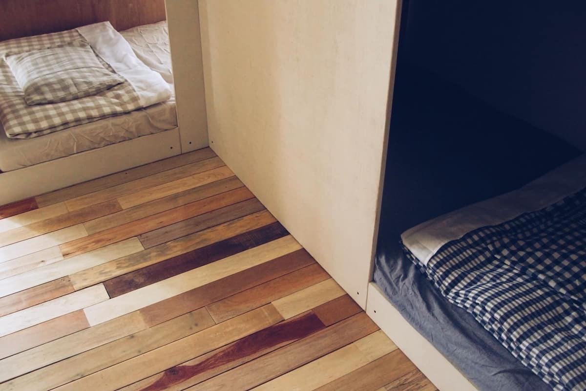 Una habitación con piso de madera, una litera con manta a cuadros y una pared lisa.