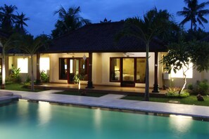 A resort with a swimming pool, palm trees, and a well-lit building with large windows. - Hotel Melamun (Buleleng)