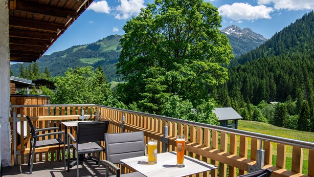 Une terrasse en bois avec des sièges d’extérieur et deux verres de bière posés sur une table, offrant une vue sur un paysage montagneux.