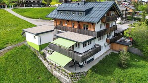 Une maison à plusieurs étages, dotée d’un toit en pente, d’une façade en bois et d’un balcon aménagé avec des sièges d’extérieur.