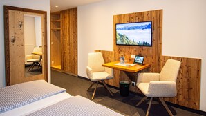 Une chambre d’hôtel avec un mur en bois, un téléviseur fixé au mur, un bureau avec un ordinateur portable, deux chaises, un lit et un plafond en bois.