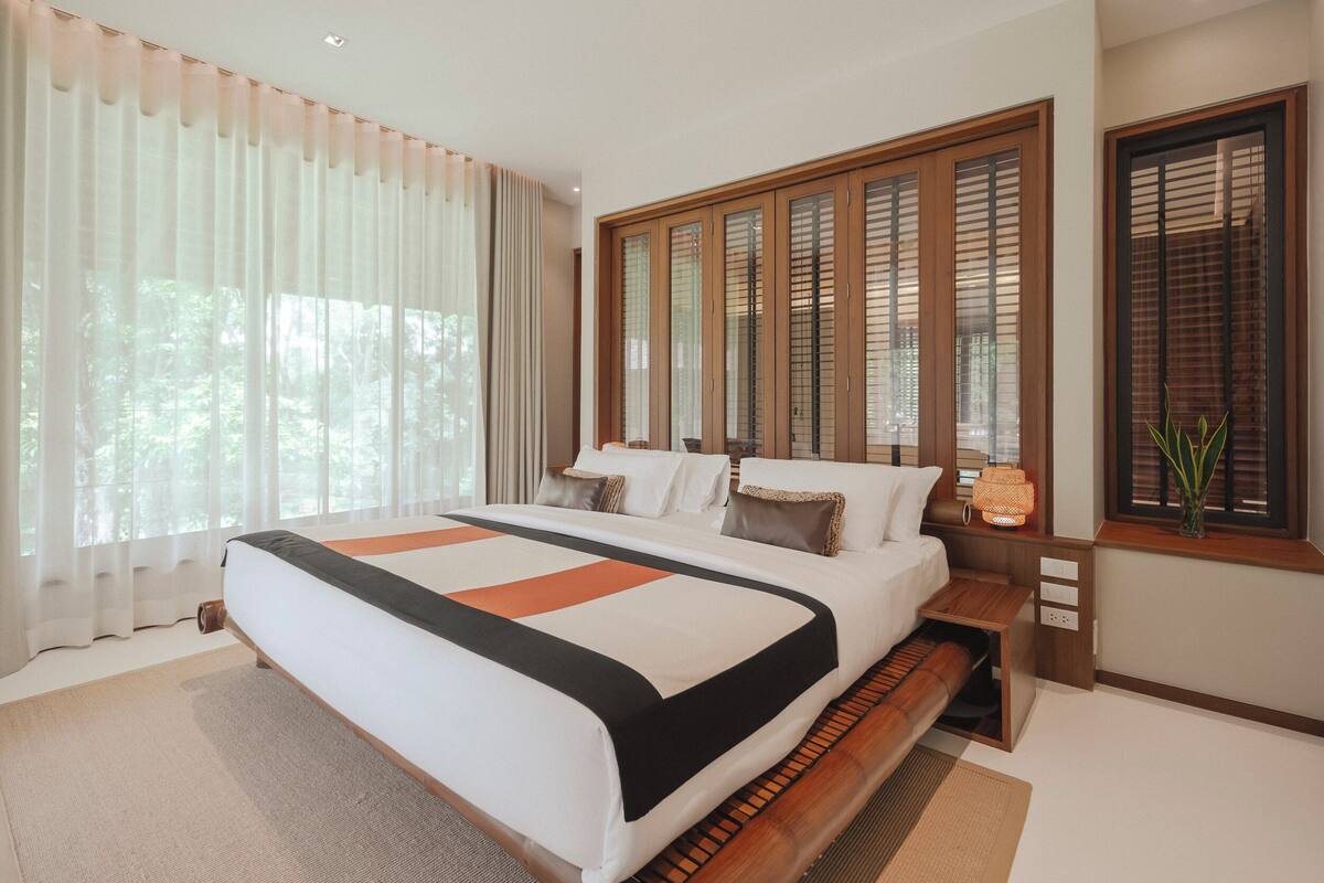 A modern bedroom with a large bed, wooden headboard, and a window with blinds.