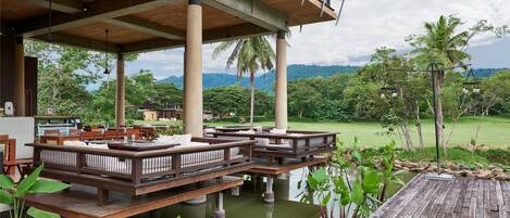 Una terraza de madera con asientos sobre el agua, sostenida por pilares, y vista a un paisaje exuberante con árboles y una cadena montañosa distante.