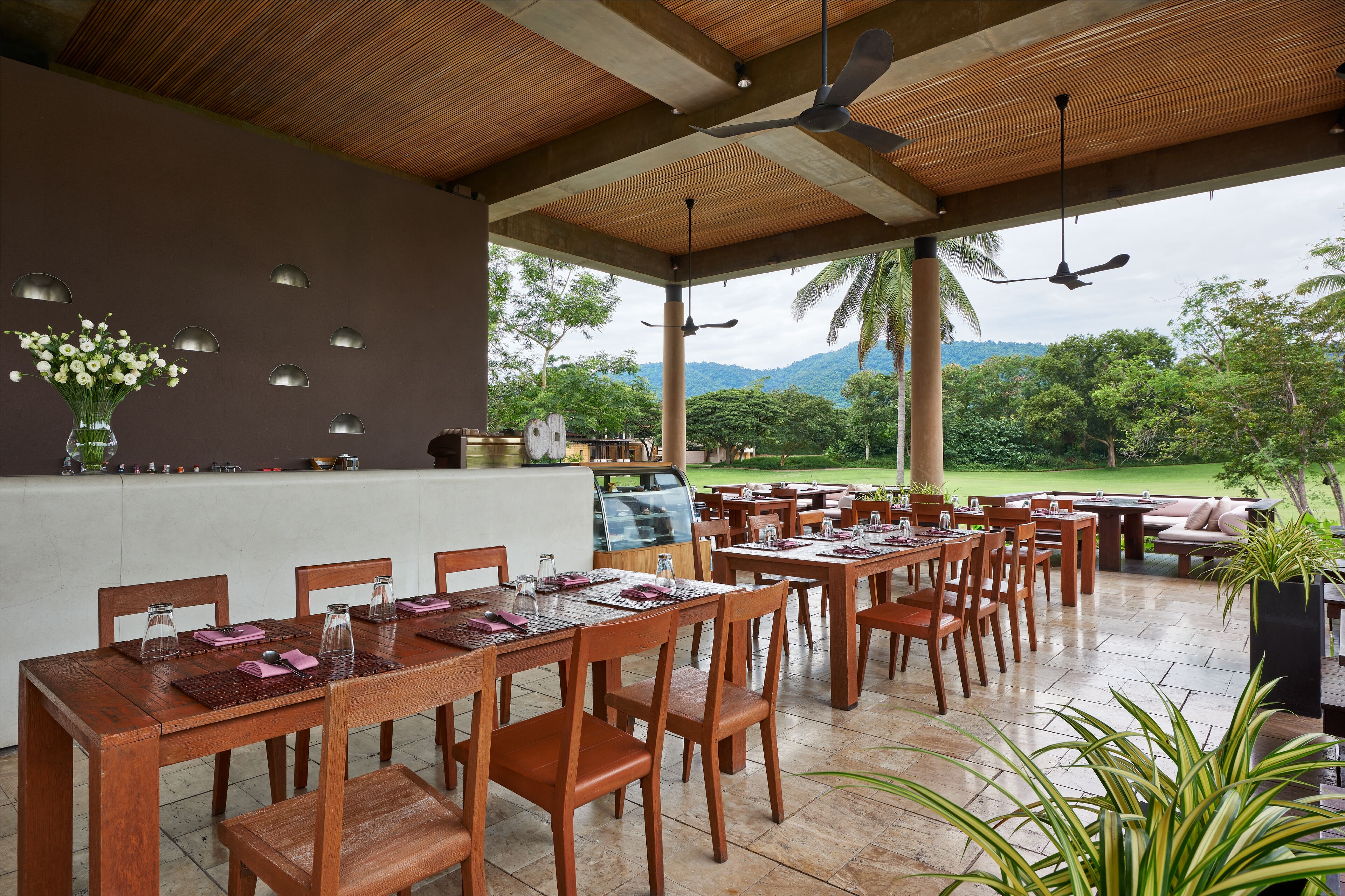 Un área de comedor al aire libre con mesas y sillas de madera, una zona de bar con mostrador e iluminación decorativa en la pared, y una vista de vegetación exuberante y montañas al fondo.