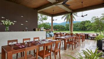Un área de comedor al aire libre con mesas y sillas de madera, una zona de bar con mostrador e iluminación decorativa en la pared, y una vista de vegetación exuberante y montañas al fondo.