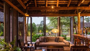 A cozy indoor seating area with a sofa, chairs, and a dining table, surrounded by large windows and plants. - Kirimaya Golf Resort Spa (Pak Chong)