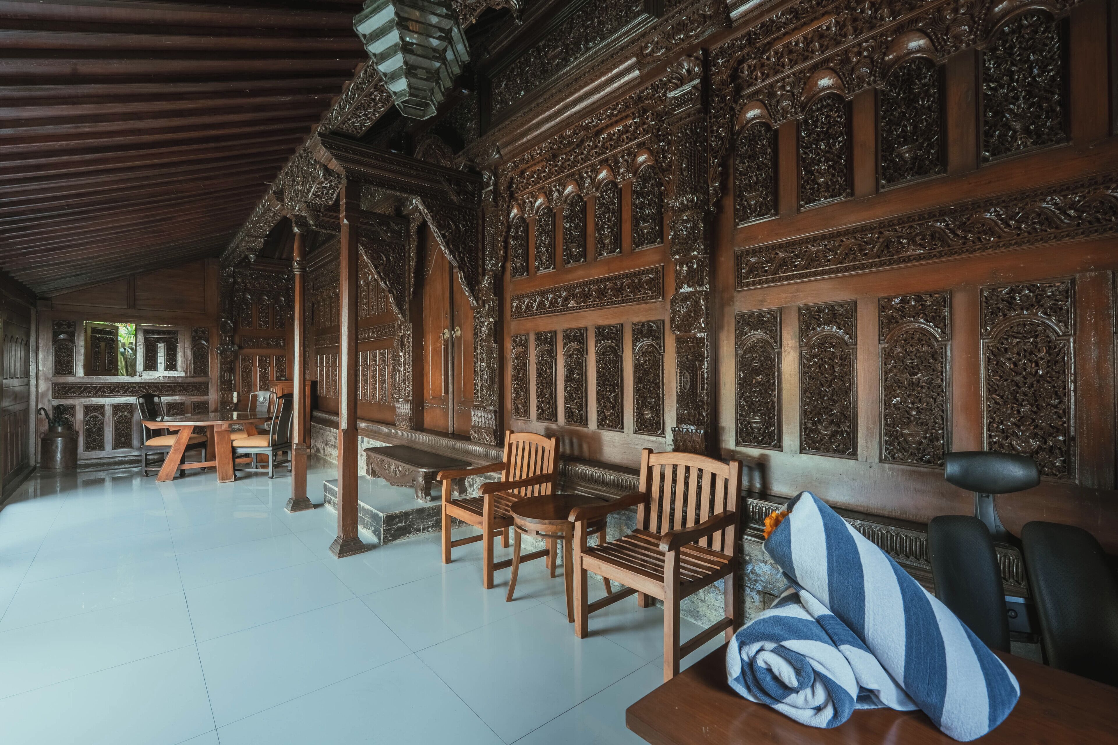 A traditional wooden interior with ornate carvings, a tiled floor, and a seating area with chairs and a bench.