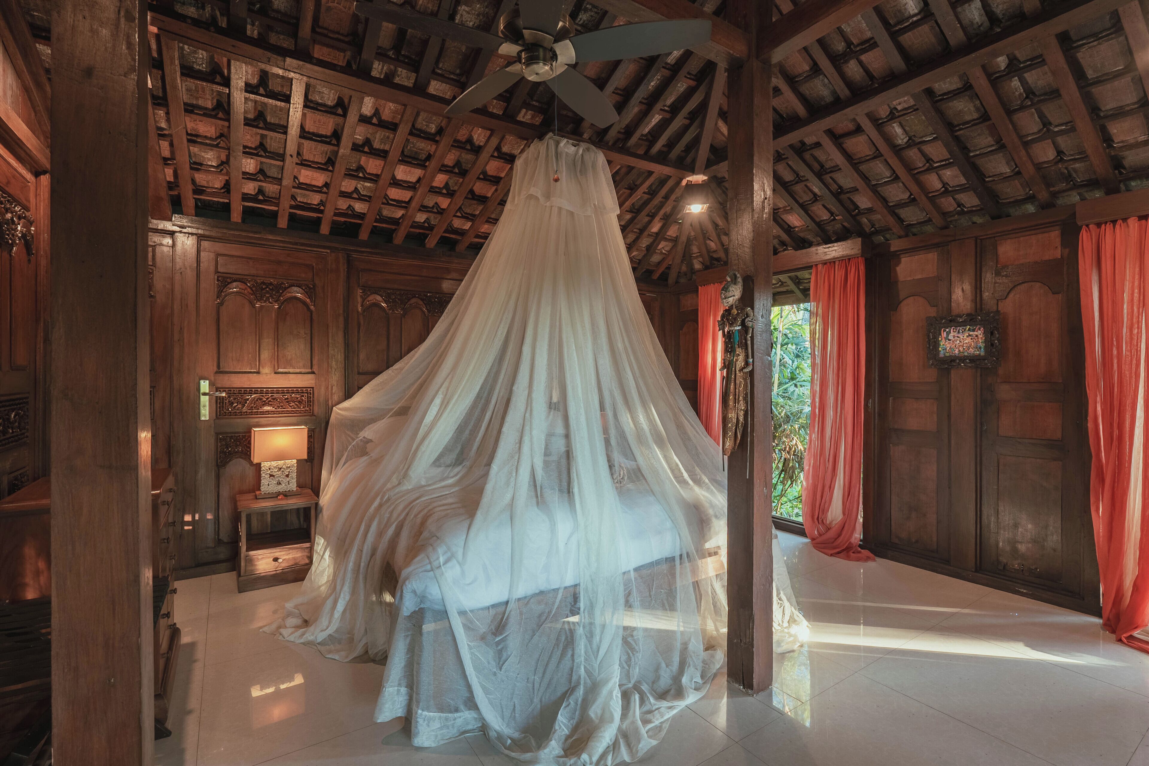 A room with a canopy bed, wooden walls, a ceiling fan, and a nightstand with a lamp.