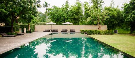 A rectangular swimming pool with a wooden deck and lounge chairs, surrounded by greenery and a low wall.