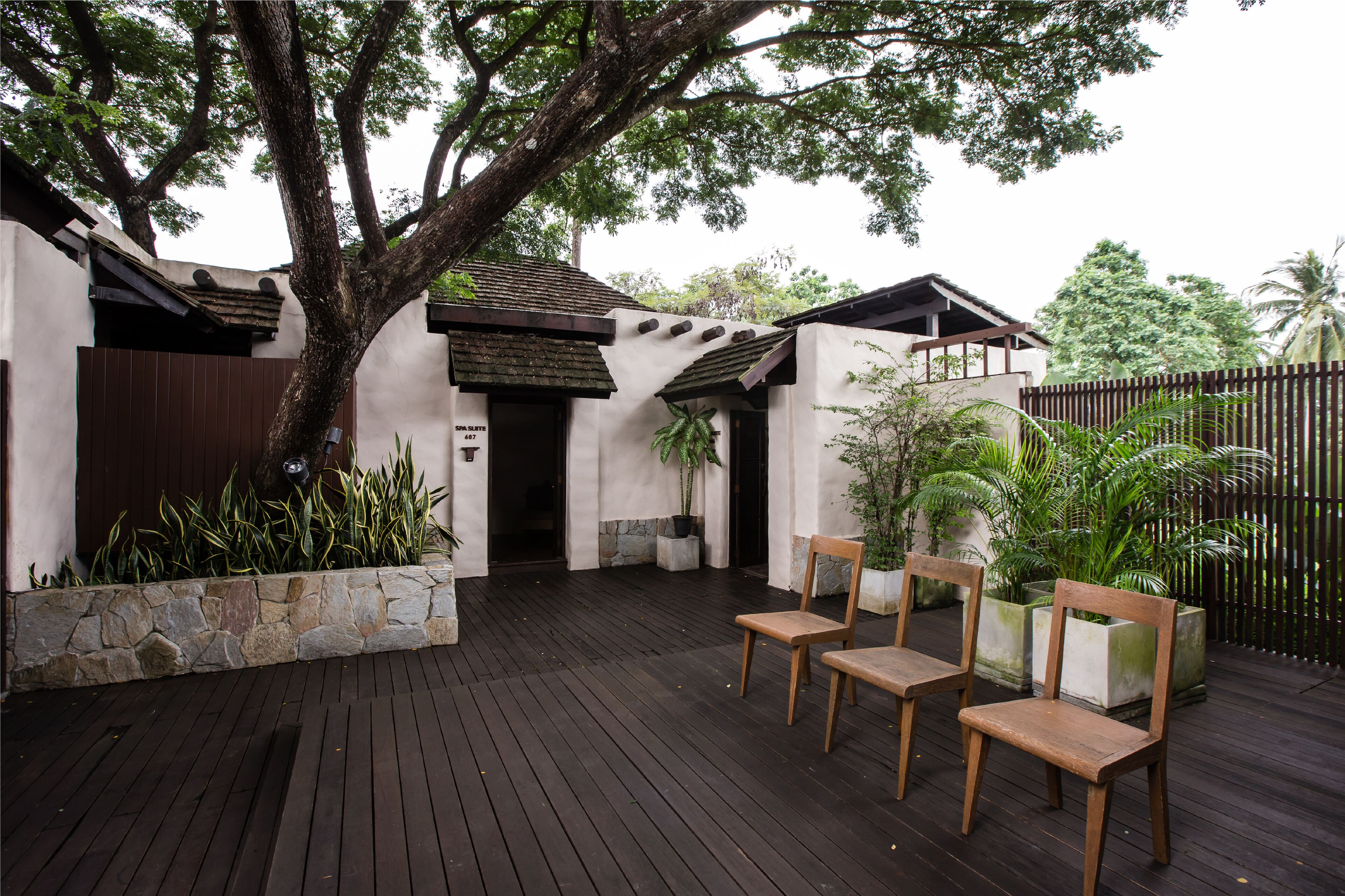 A wooden deck with chairs and potted plants, a stone wall, and a building with a tiled roof.