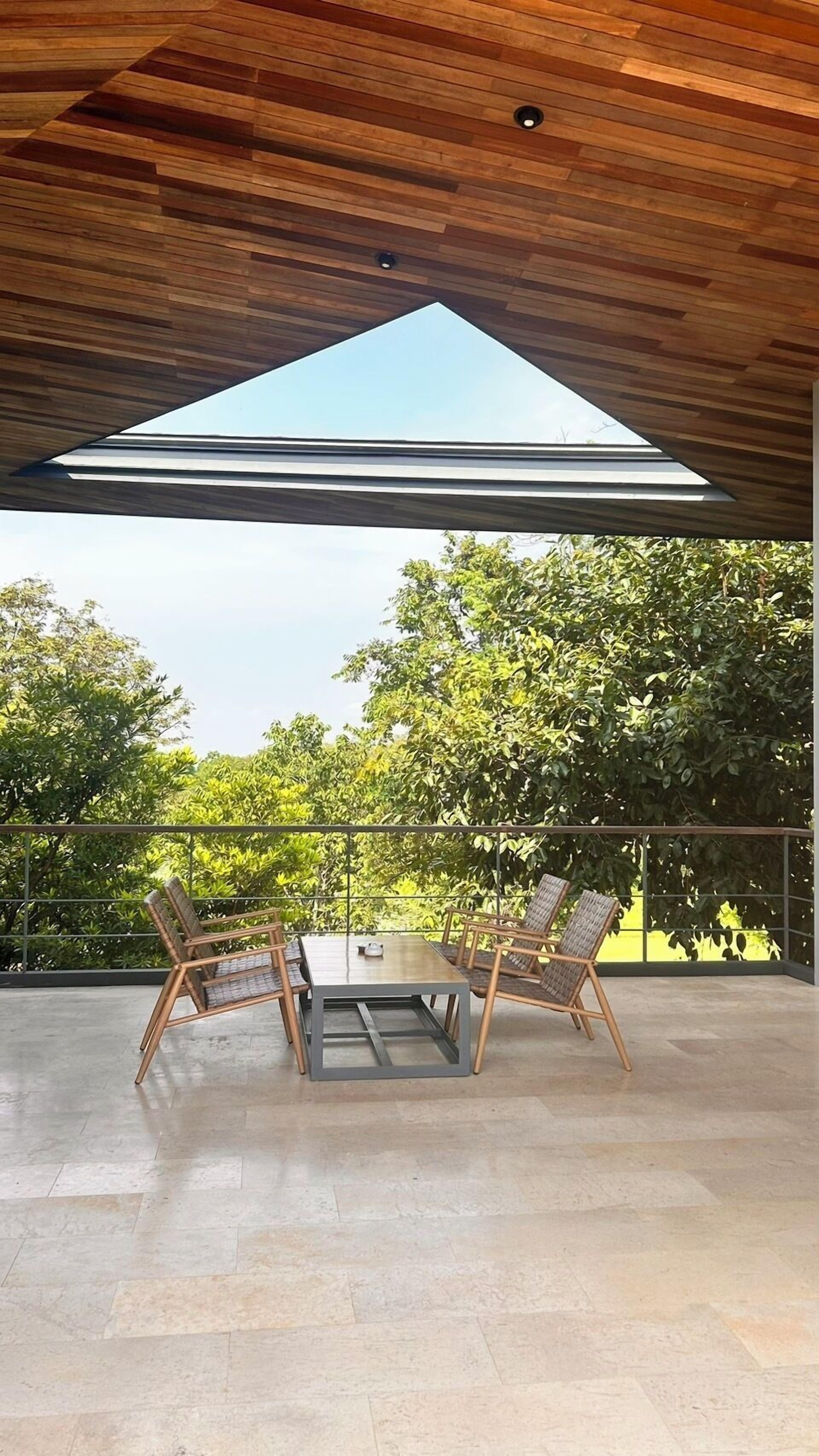 Een overdekt terras met een tafel en stoelen, een uitzicht op bomen en een houten plafond.