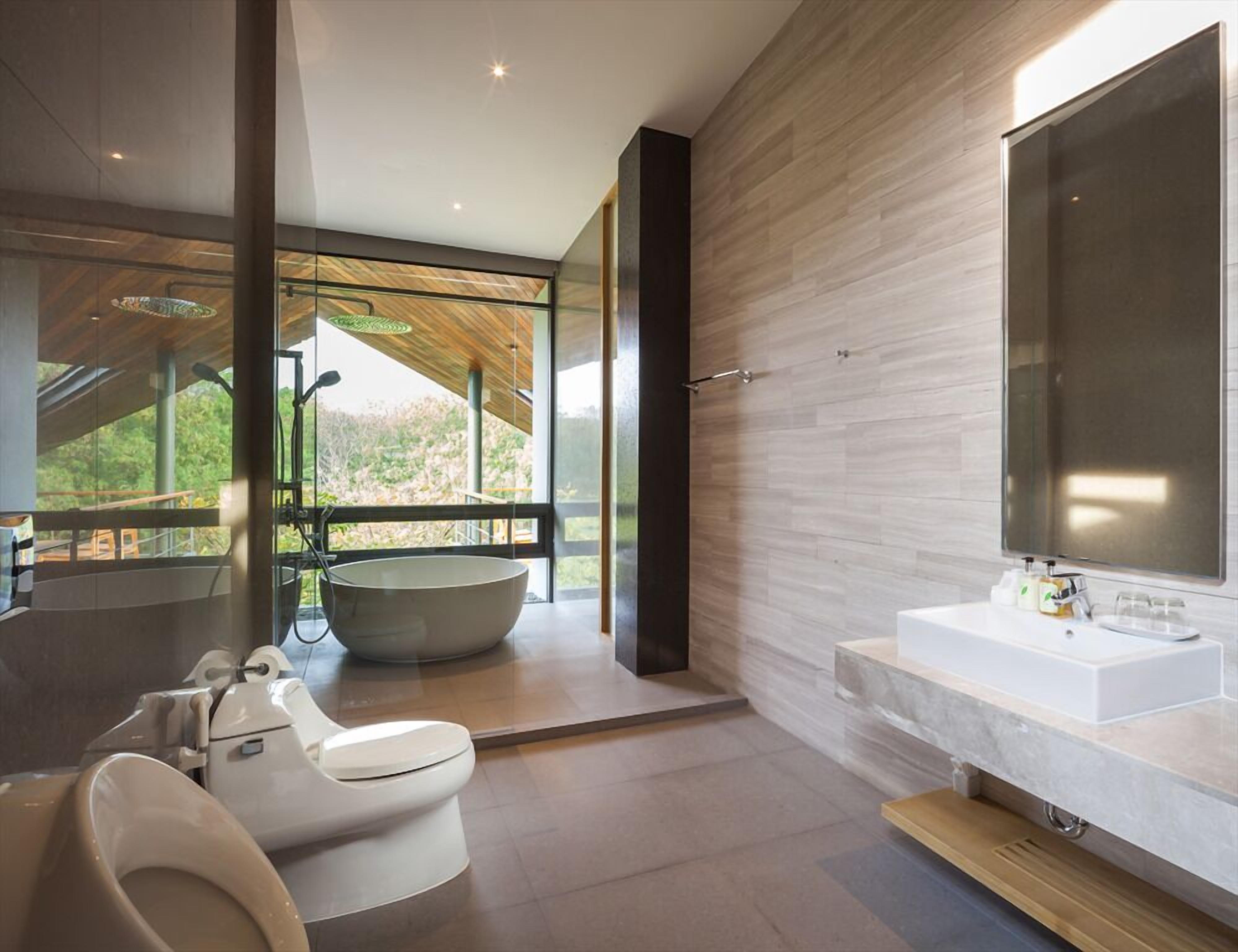 Ein modernes Badezimmer mit Badewanne, Toilette und Waschbecken, großen Fenstern und Blick auf Grünflächen.