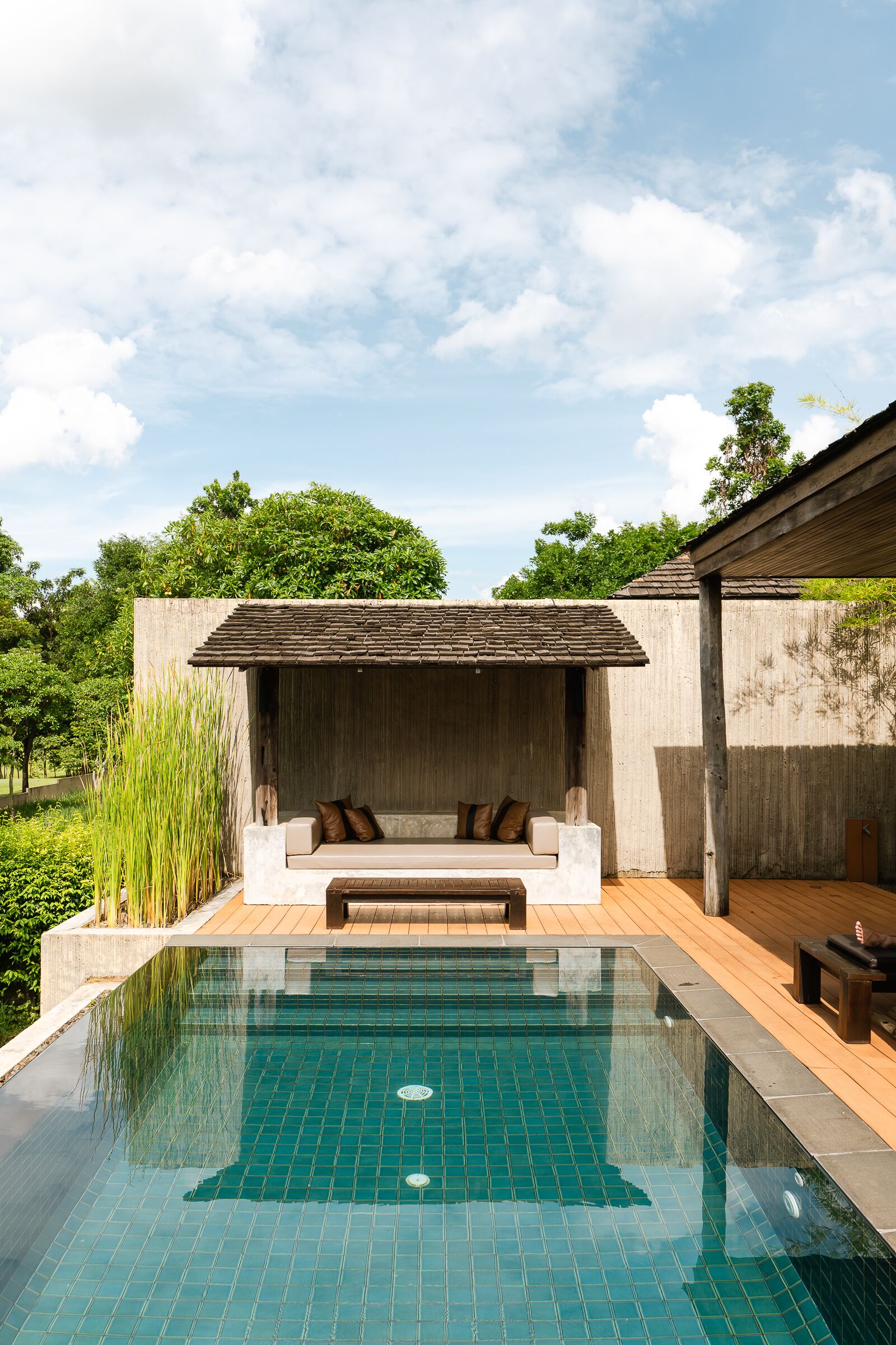 A swimming pool with a wooden deck, a concrete wall with a built-in seating area, and a view of greenery in the background.