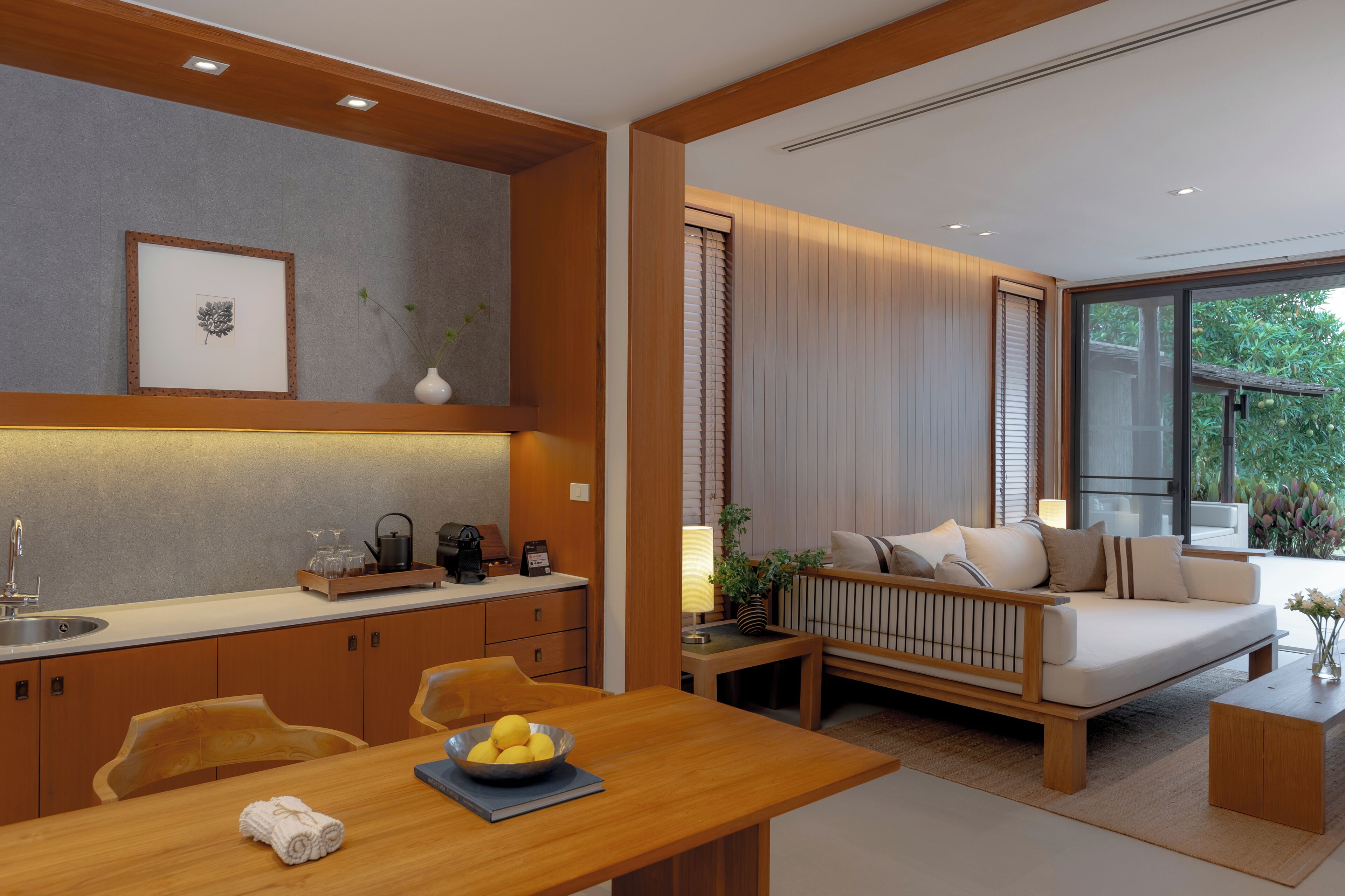 A modern kitchen with a dining area, a sofa, and a view of greenery.