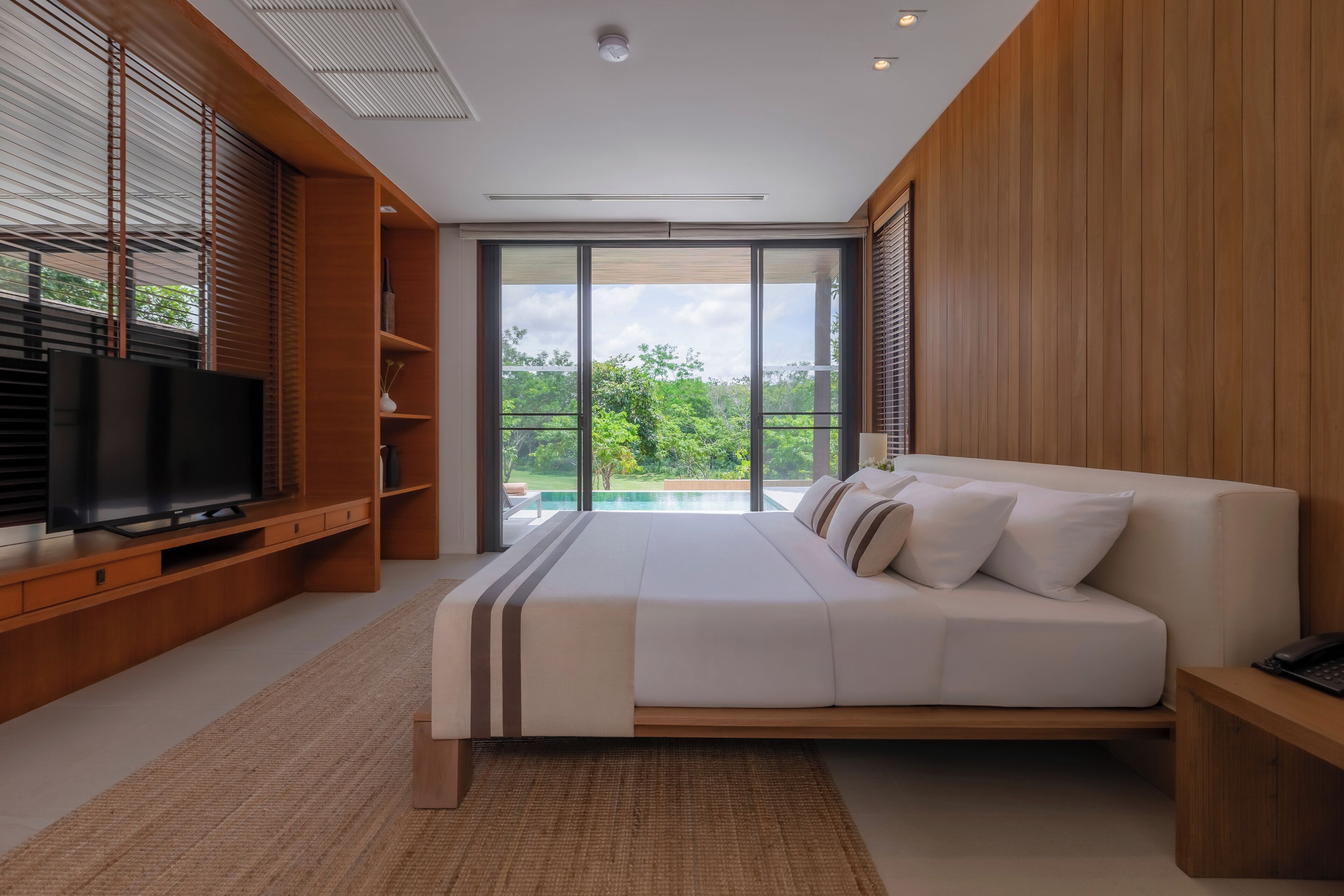 Ein modernes Schlafzimmer mit einem großen Bett, einem Fernseher, Holzvertäfelung und Blick auf Grünflächen.