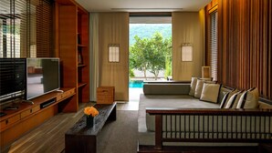 A modern living room with a flat-screen TV, wooden furniture, a sofa with cushions, a coffee table with flowers, and a view of a pool and greenery. - MUTHI MAYA Forest Pool Villa Resort (Pak Chong)