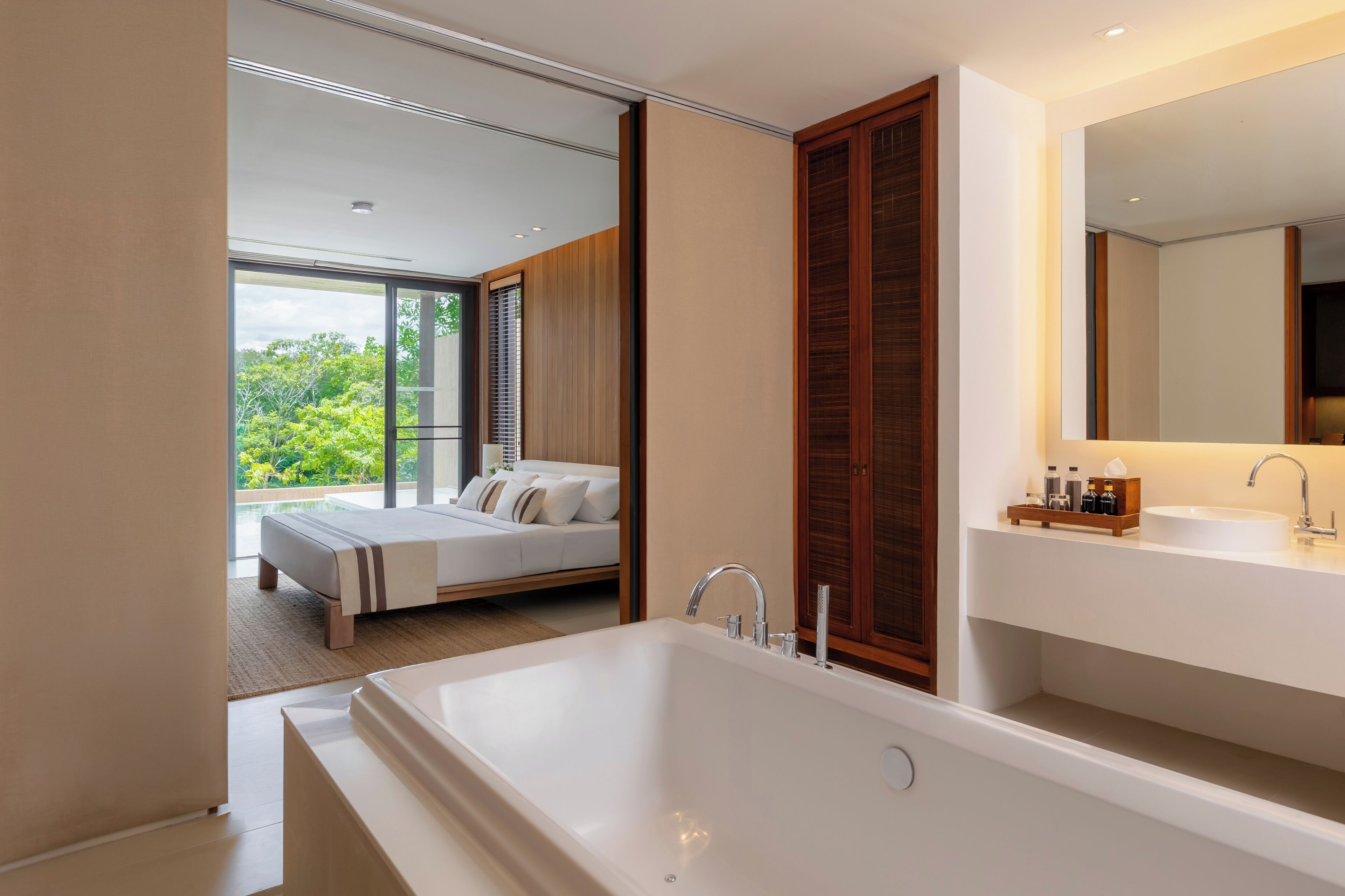 A modern bathroom with a large bathtub, a vanity with a mirror, and a bed visible through a glass door.
