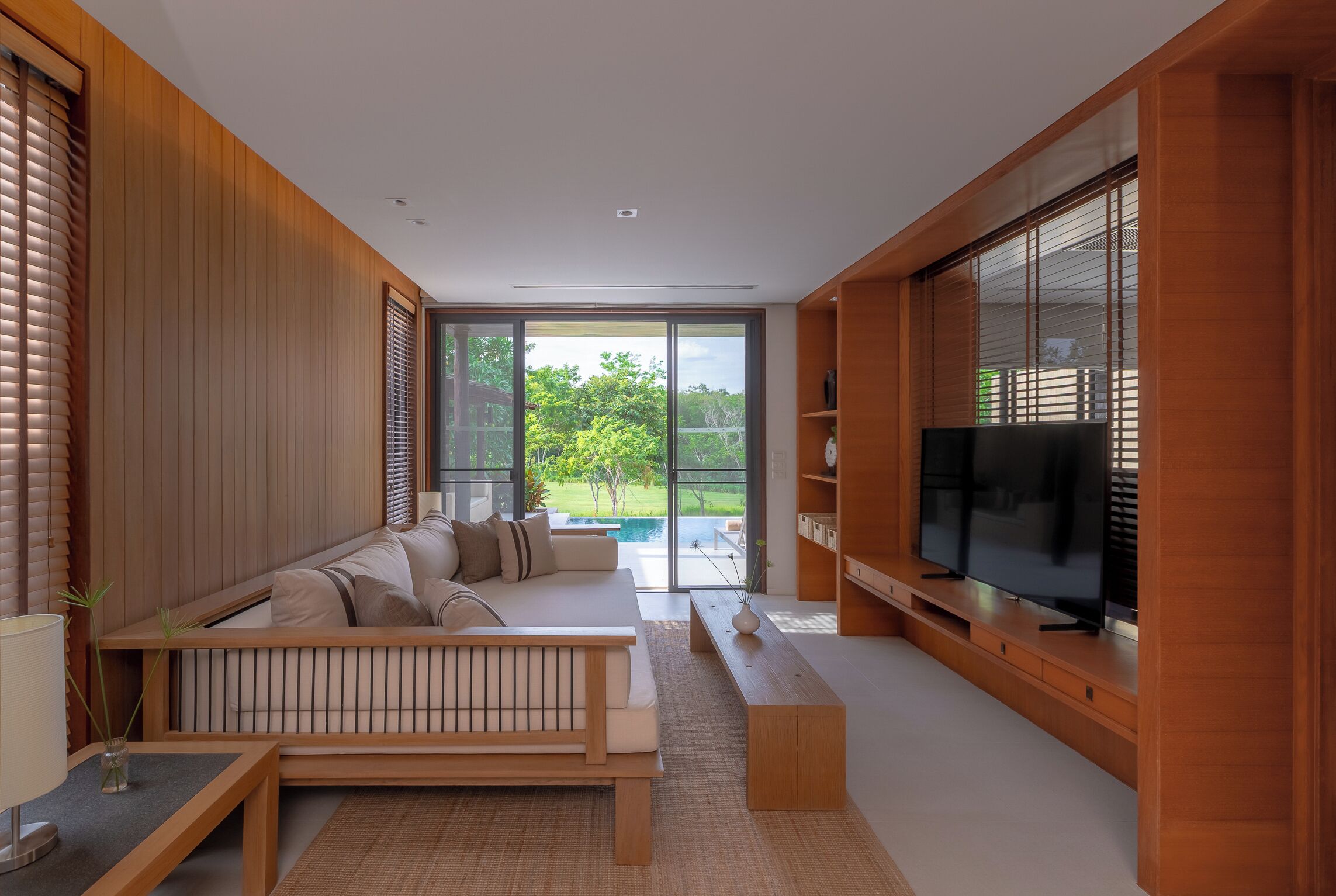 A modern living room with a sectional sofa, a flat-screen TV, and large windows overlooking a pool.
