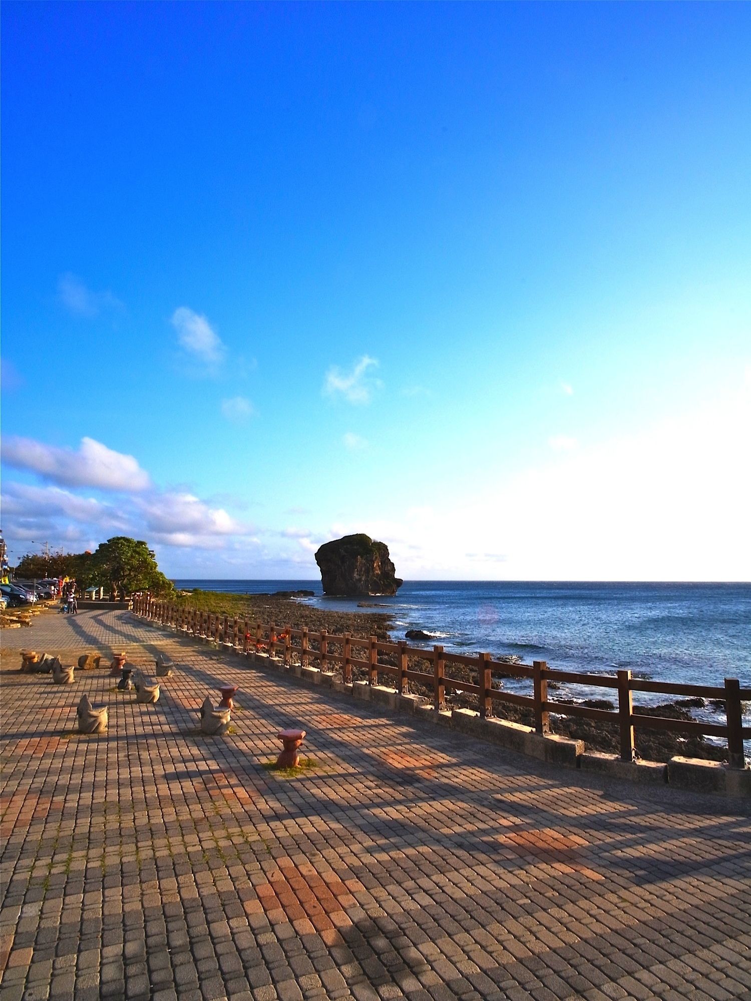 A paved walkway with a wooden fence, leading to a rocky coastline with a prominent rock formation.