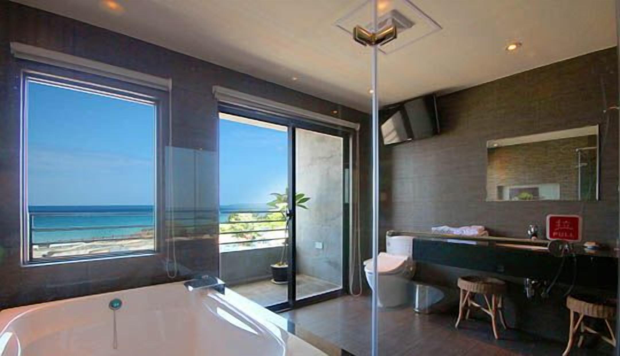 A modern bathroom with a large bathtub, a toilet, and a sink. There is a sliding glass door leading to a balcony with a view of the beach.