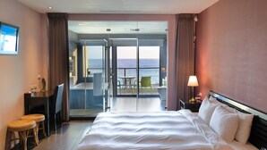 Habitación de hotel con una cama grande, un escritorio, una silla, una mesa con sillas y vistas al mar a través de una puerta de cristal.
