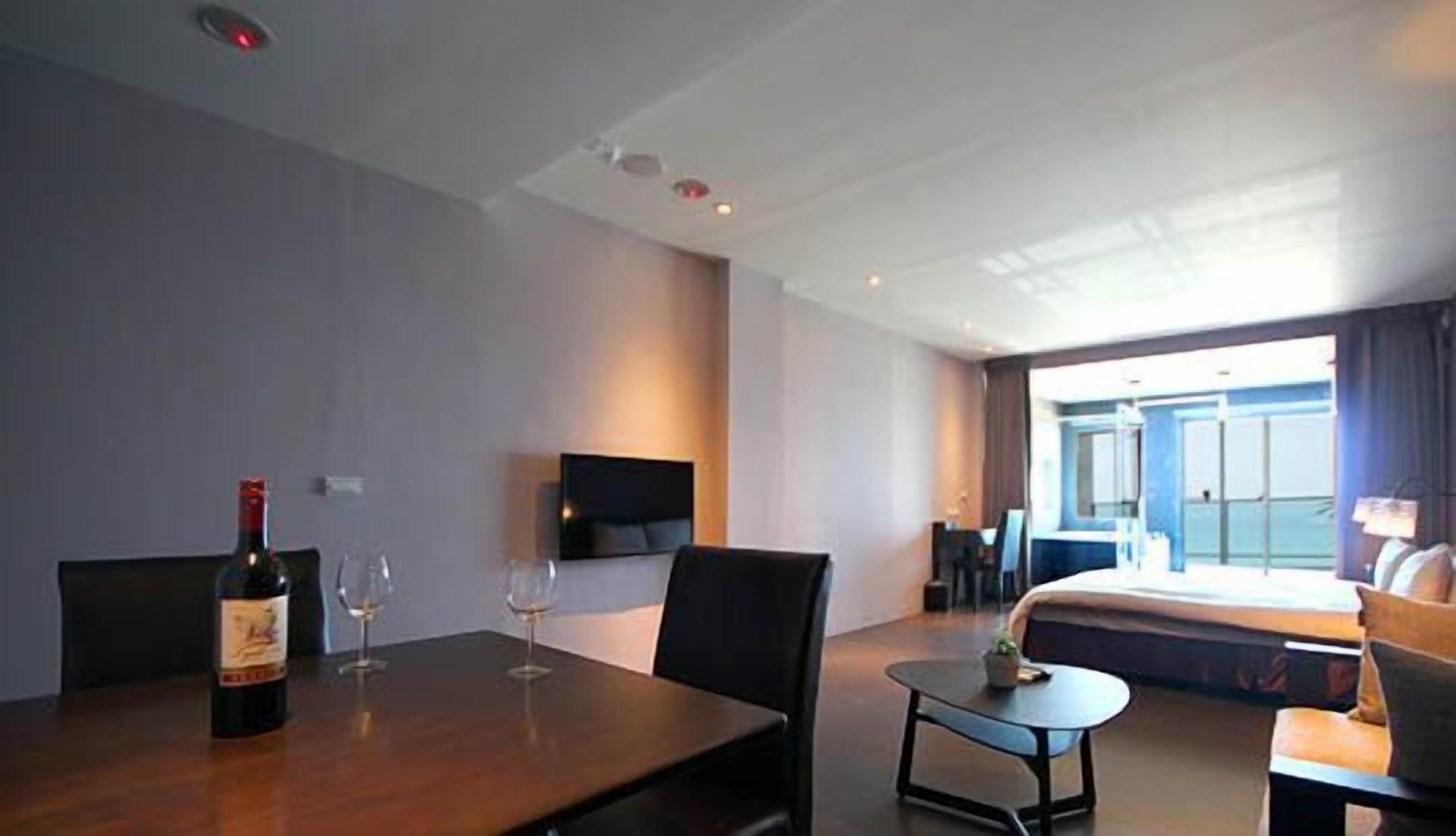 A hotel room with a bed, a desk, a chair, a small table, and a bottle of wine.