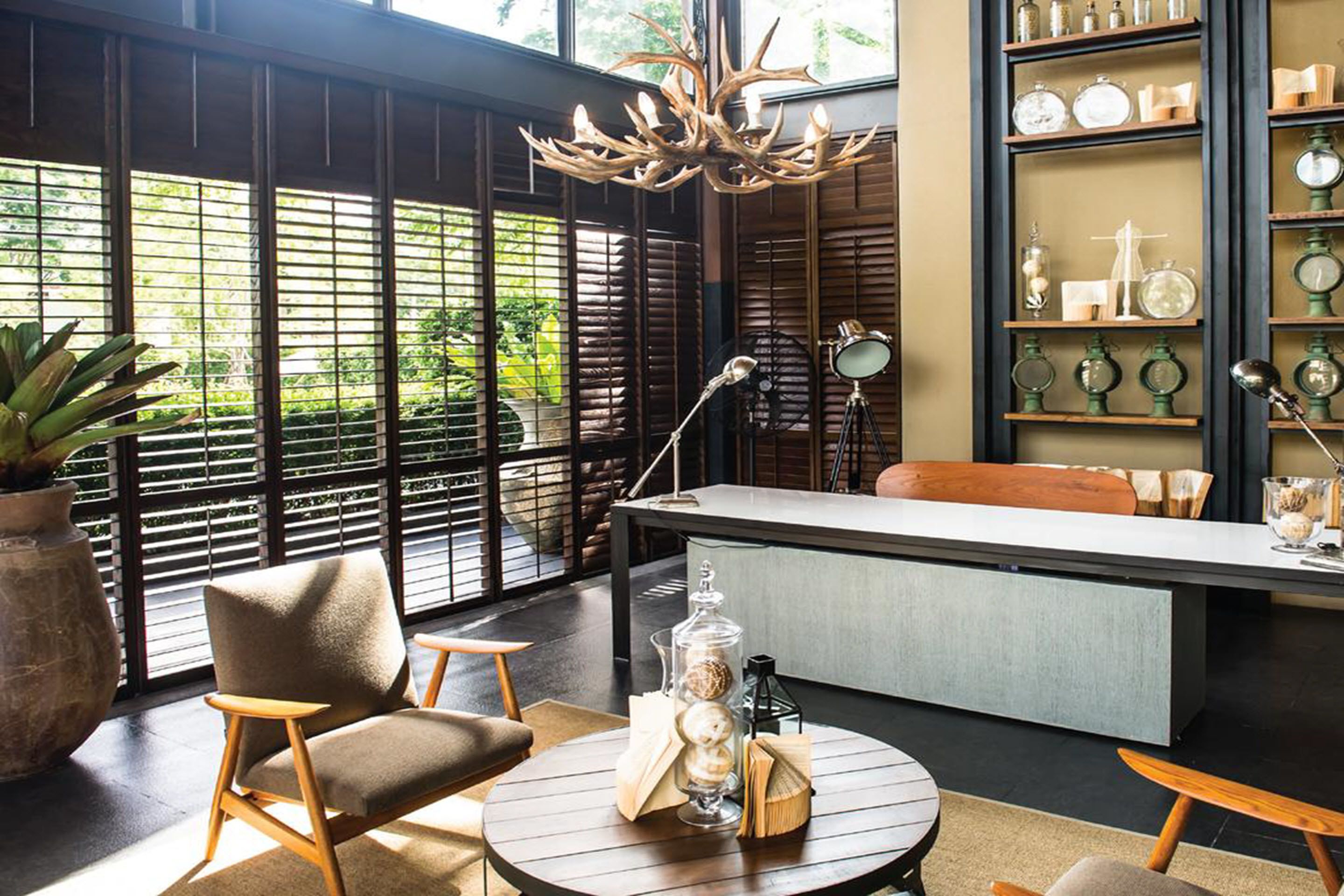 Un interior moderno con zona de comedor, un ventanal con persianas, una araña y estanterías con objetos decorativos.