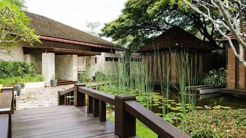 A wooden deck leading to a building with a tiled roof, surrounded by lush greenery and a pond with lily pads.